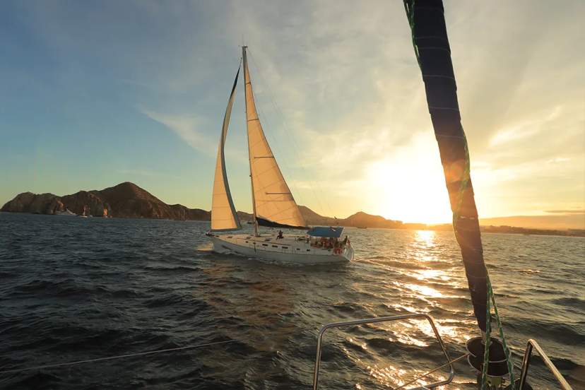 Sailboat glides over the sea at sunset, with rocky cliffs in the distance.