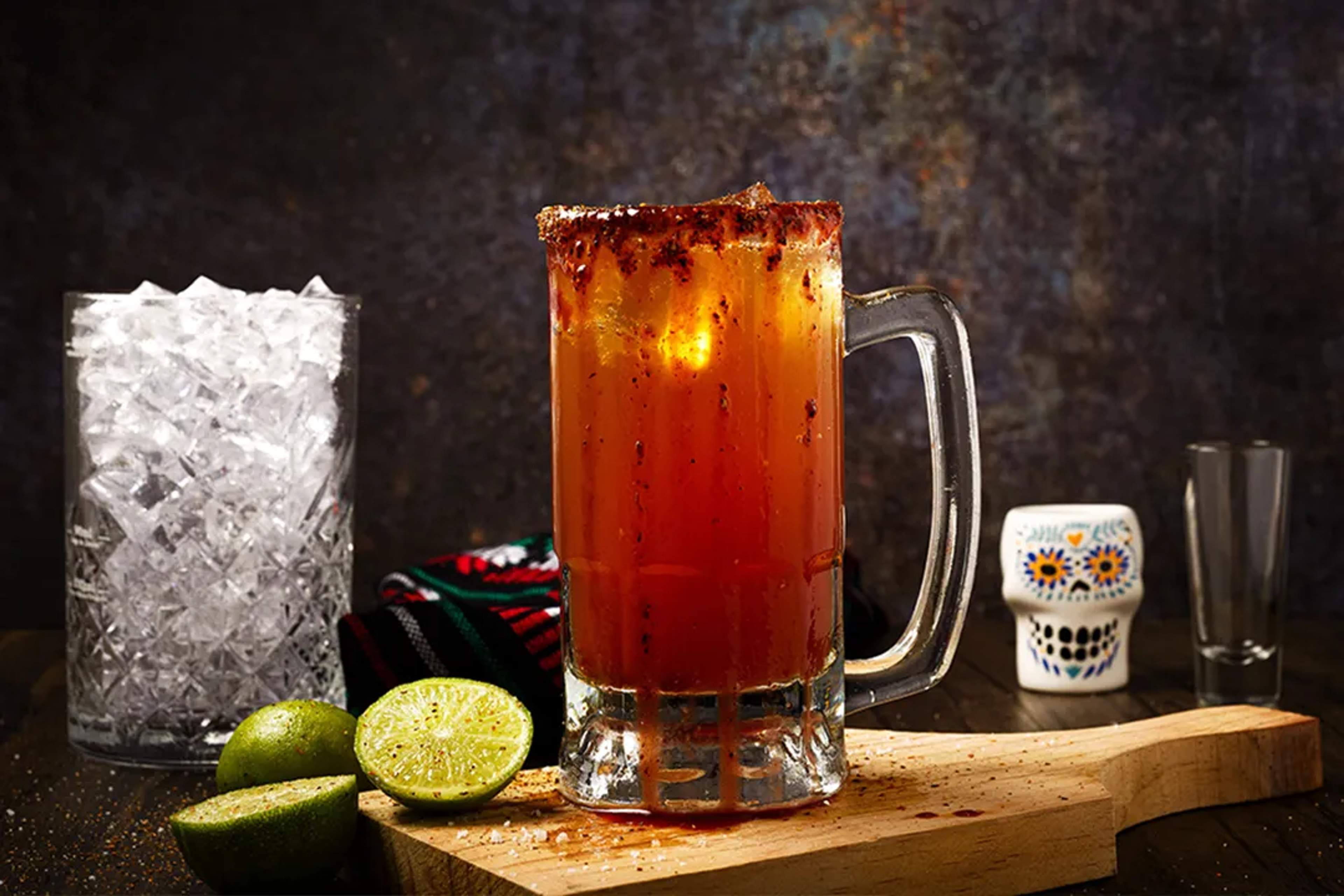 Refreshing michelada in a frosty mug with chili rim, lime, and ice on a rustic wooden board.