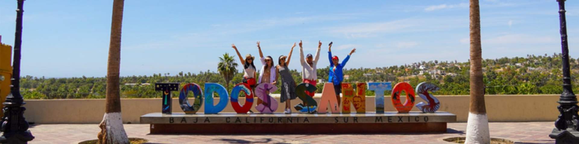 A group of people pose with arms raised behind a colorful "Todos Santos" sign in Baja California Sur, Mexico, with palm trees in the background.