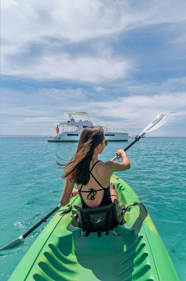 Mujer navegando en kayak por las aguas turquesa de Balandra, con un catamarán de Cabo Adventures al fondo.