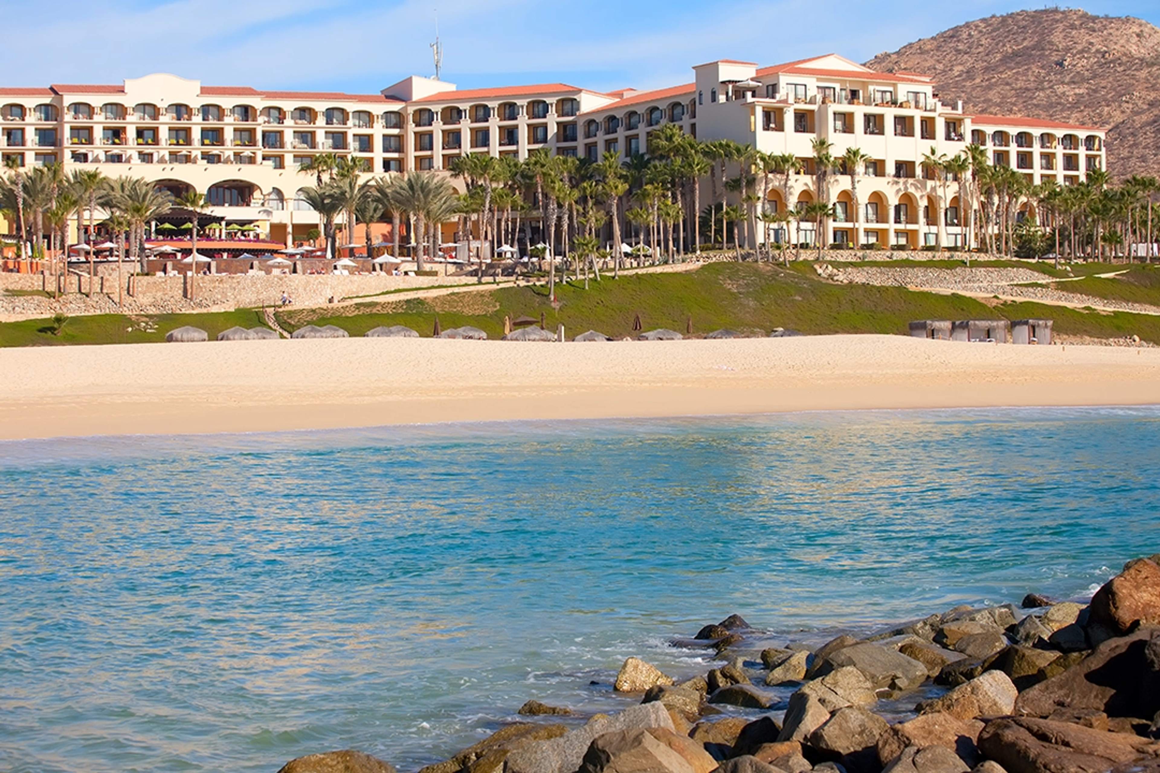 Un hotel de lujo frente al mar en Cabo San Lucas, rodeado de palmeras, arena dorada y aguas turquesas.