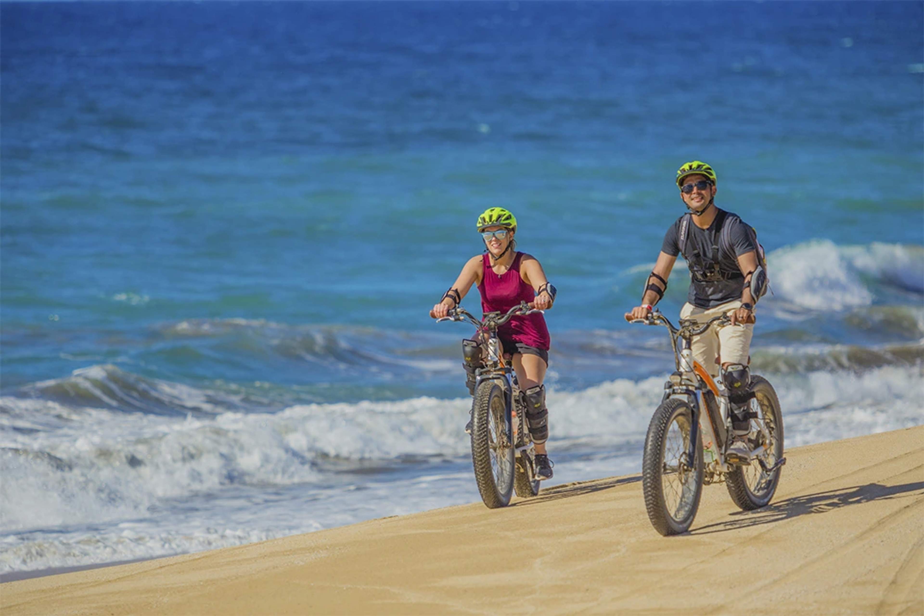 Dos personas andando en bicicleta por una playa de arena con equipo de protección, disfrutando del paisaje costero y las olas de fondo.