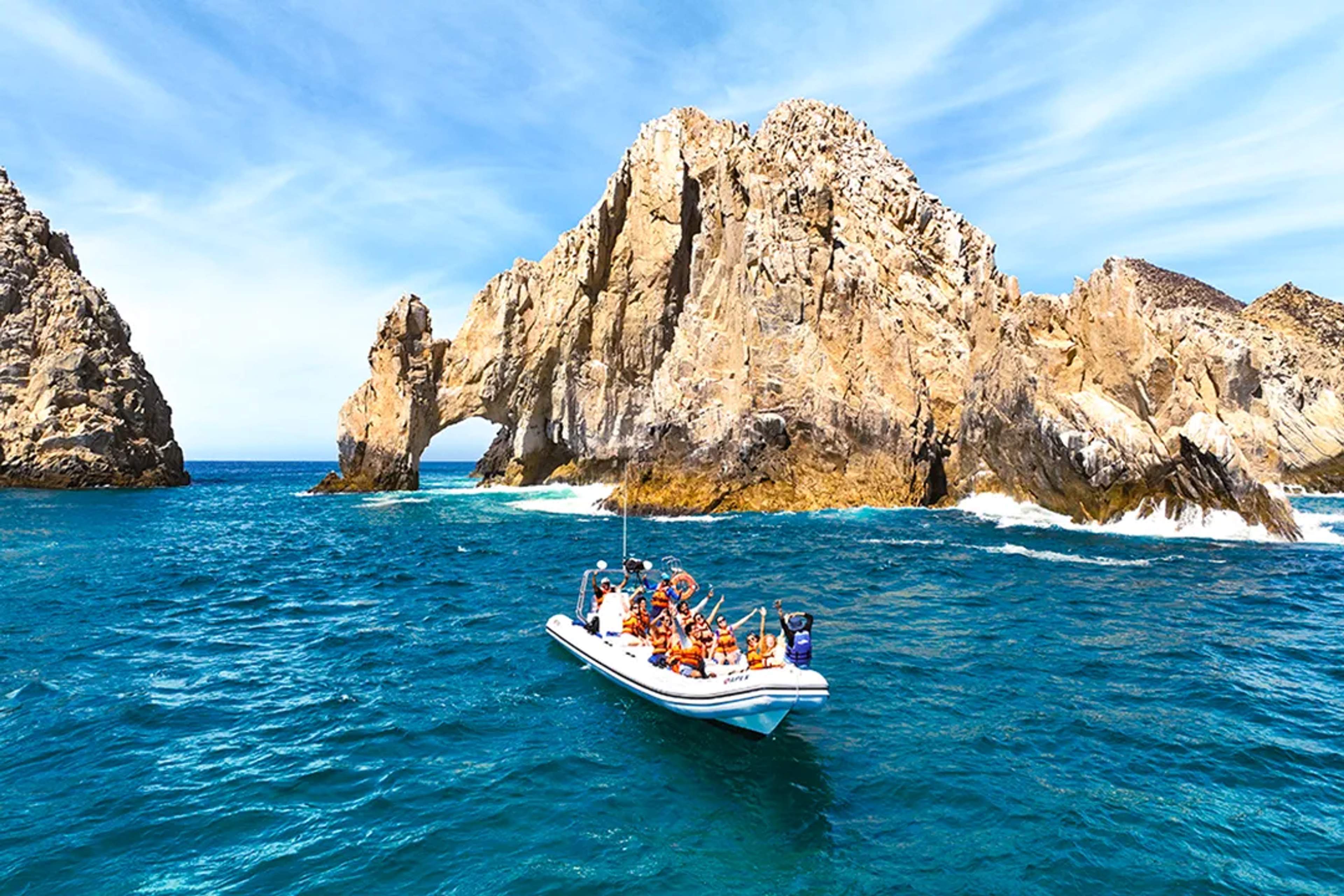 A luxury catamaran with guests enjoying the sea near a rocky coast under a bright blue sky.