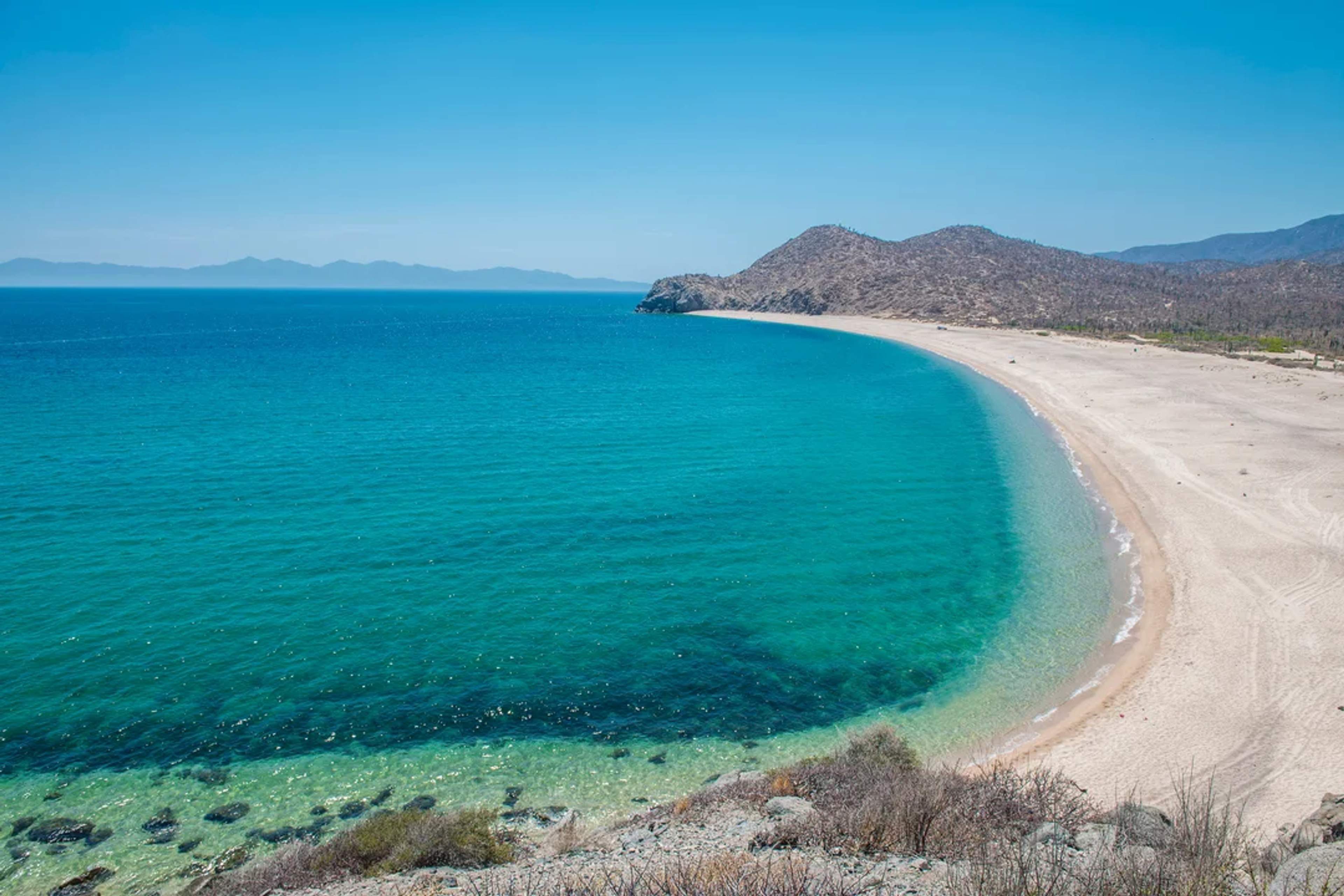 Playa aislada de arena dorada y aguas turquesa, rodeada de colinas áridas bajo un cielo despejado.