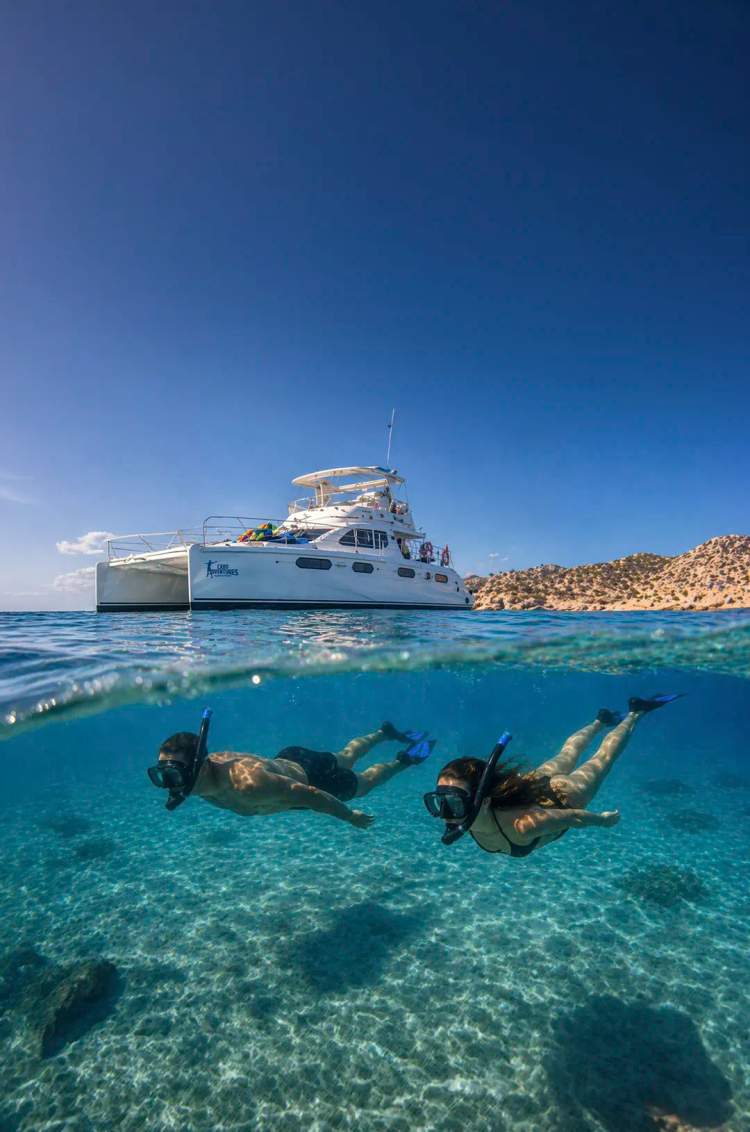 Couple snorkeling in clear turquoise waters below luxury catamaran near Cabo San Lucas desert coastline, Los Cabos Mexico