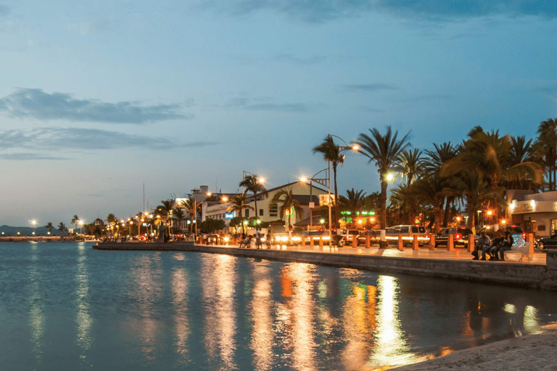 Malecón de La Paz al anochecer con luces de la ciudad reflejándose en el agua tranquila, palmeras bordeando el paseo y un cielo sereno.
