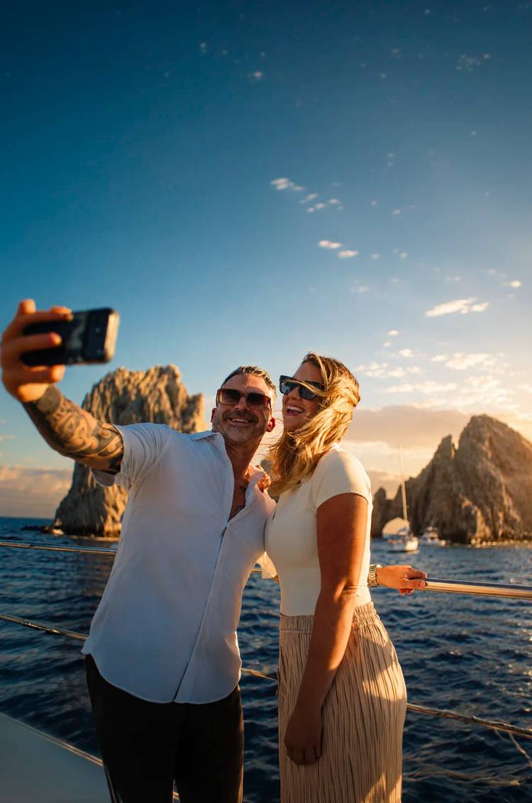 Couple taking a selfie during a sunset catamaran cruise in front of the Arch of Cabo San Lucas.