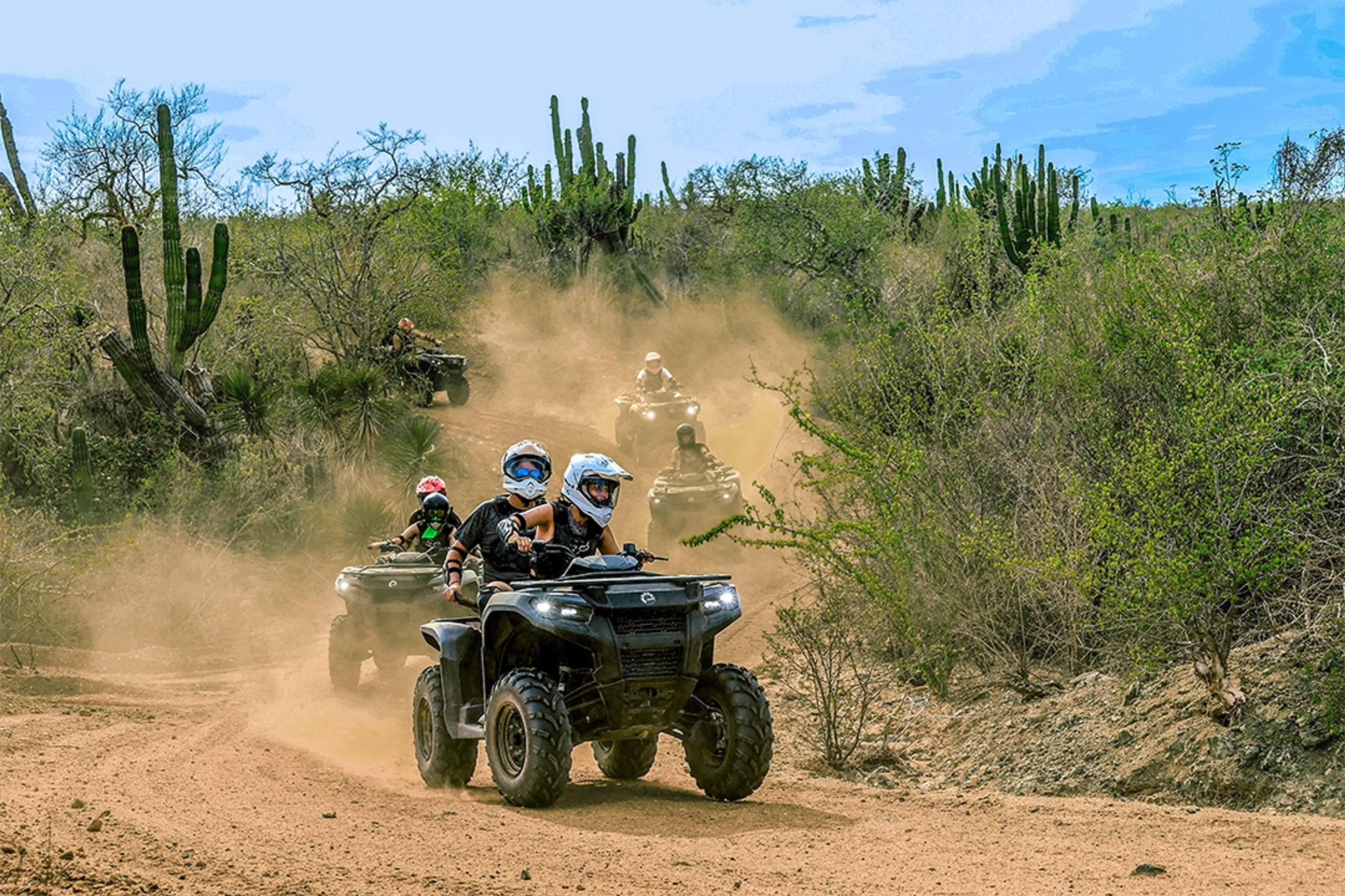 People riding ATVs on a dusty trail in the desert, surrounded by cacti and arid vegetation.