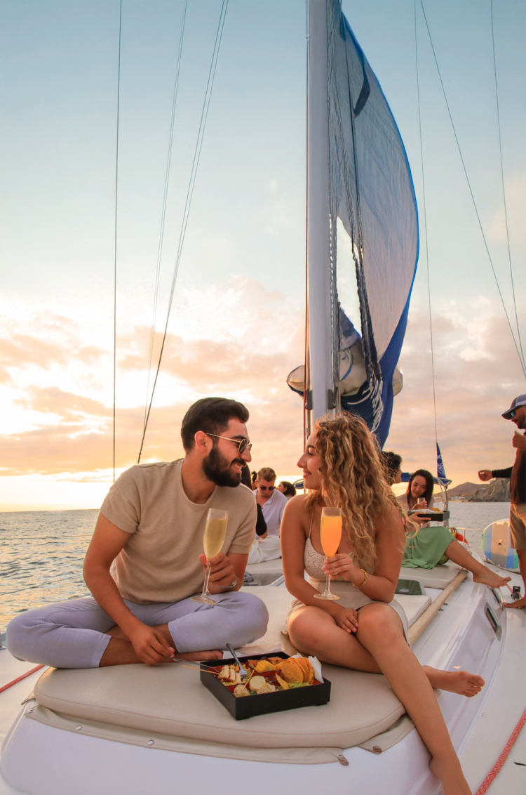 A couple enjoys drinks & snacks on a sunset cruise, smiling at each other as the ocean and sky glow warmly in the background.