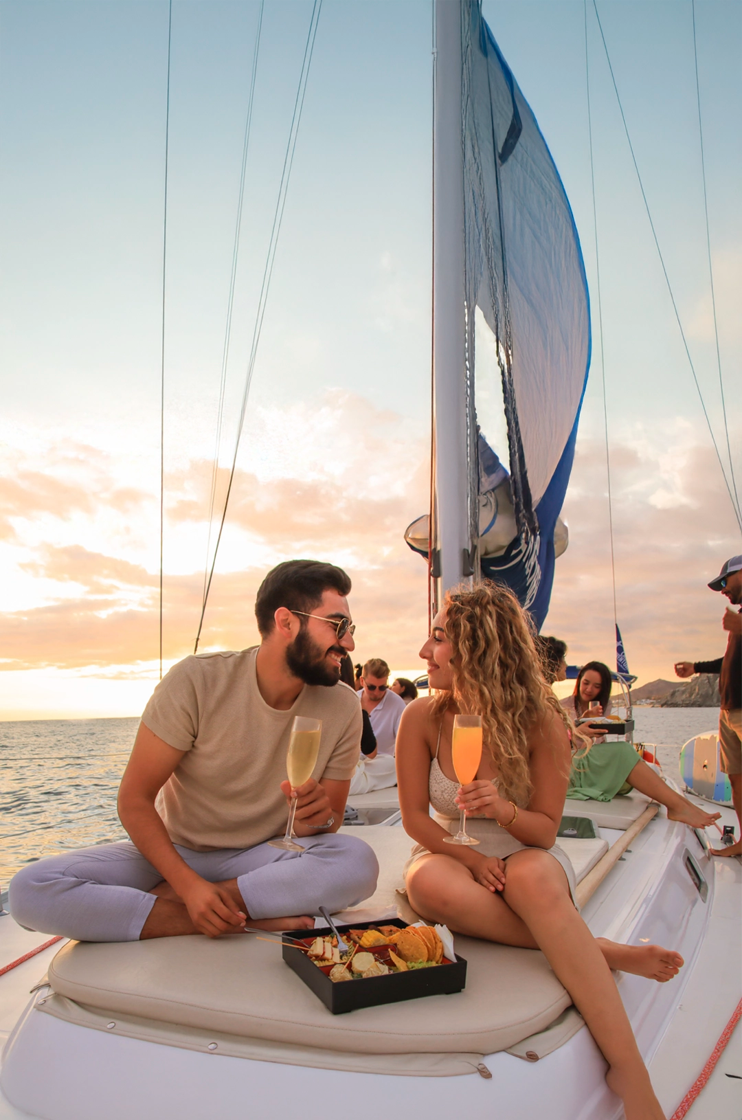 Una pareja disfruta de bebidas y aperitivos en un crucero al atardecer, con el océano y cielo cálido de fondo.