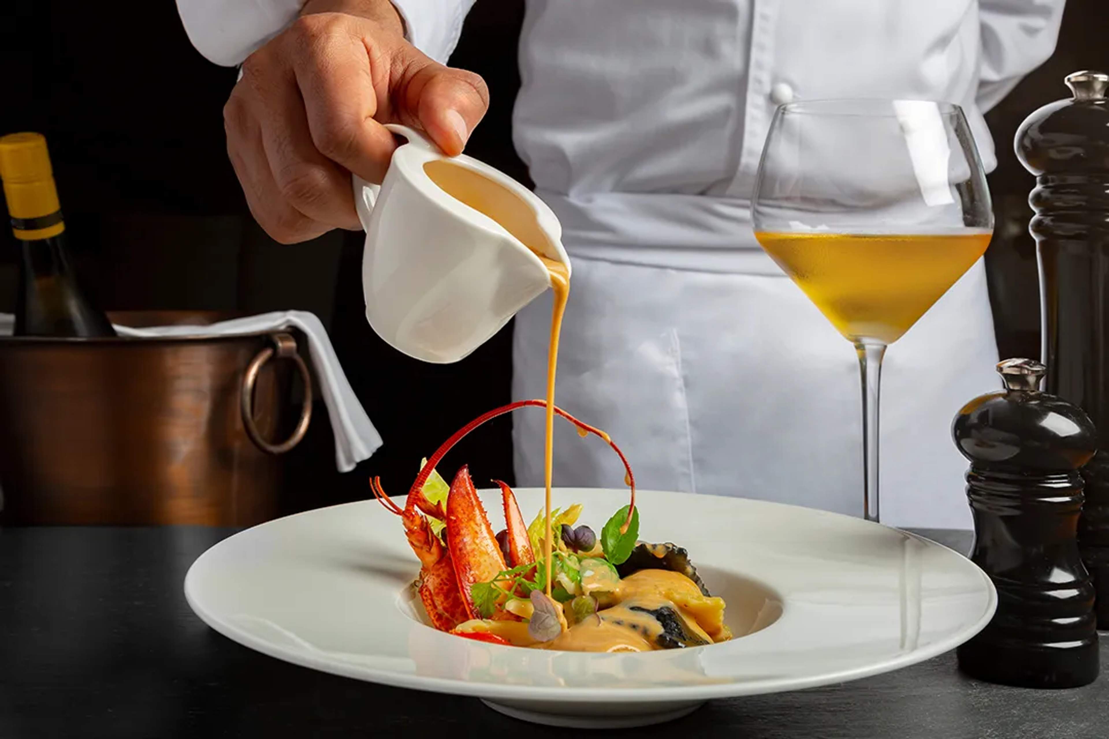 Un chef vertiendo una salsa rica sobre un plato gourmet con langosta, acompañado de una copa de vino blanco.