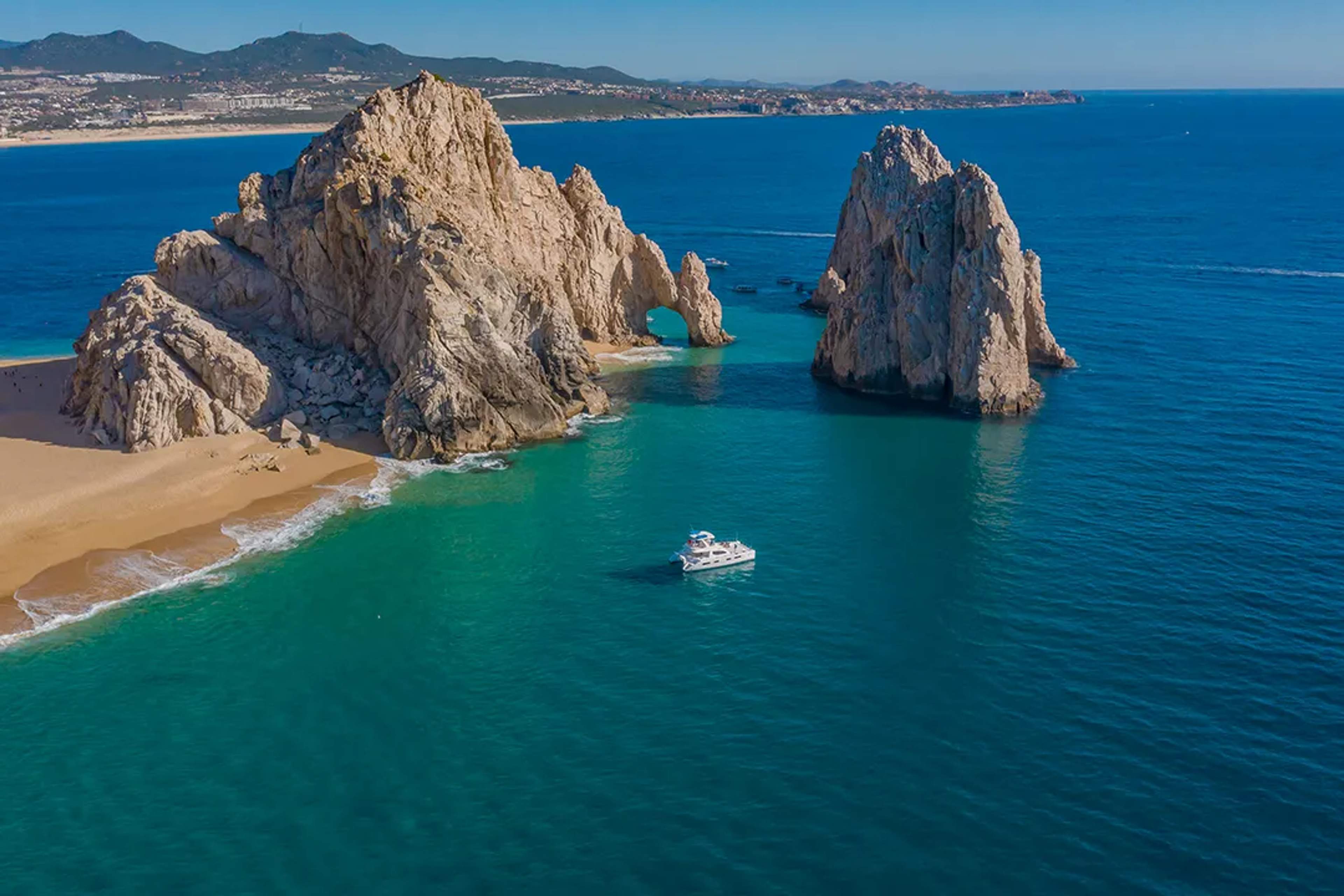 A scenic aerial view of a turquoise bay with clear shallow waters, surrounded by rocky hills and arid terrain.