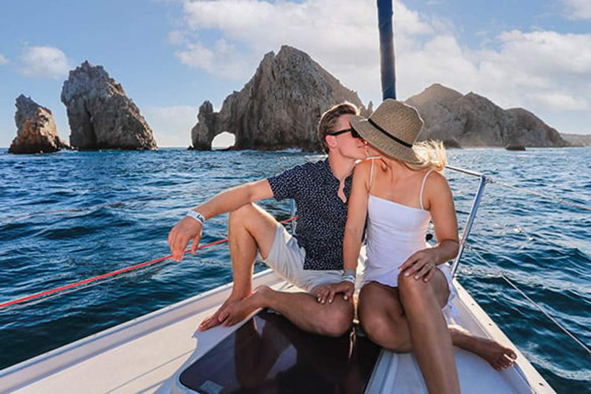 A couple kisses on a boat with the famous Land’s End Arch in the background, enjoying a romantic tour in Cabo San Lucas.