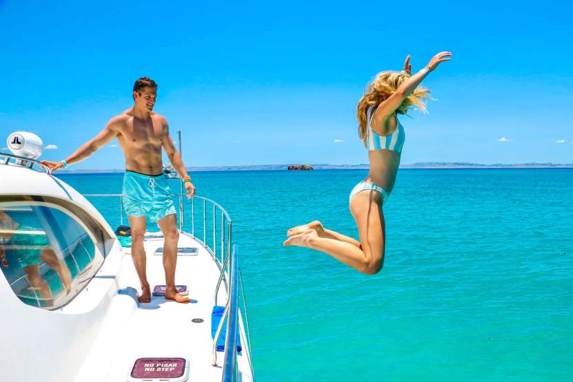 Woman jumps joyfully off a yacht into turquoise sea, while man watches under a clear blue sky.