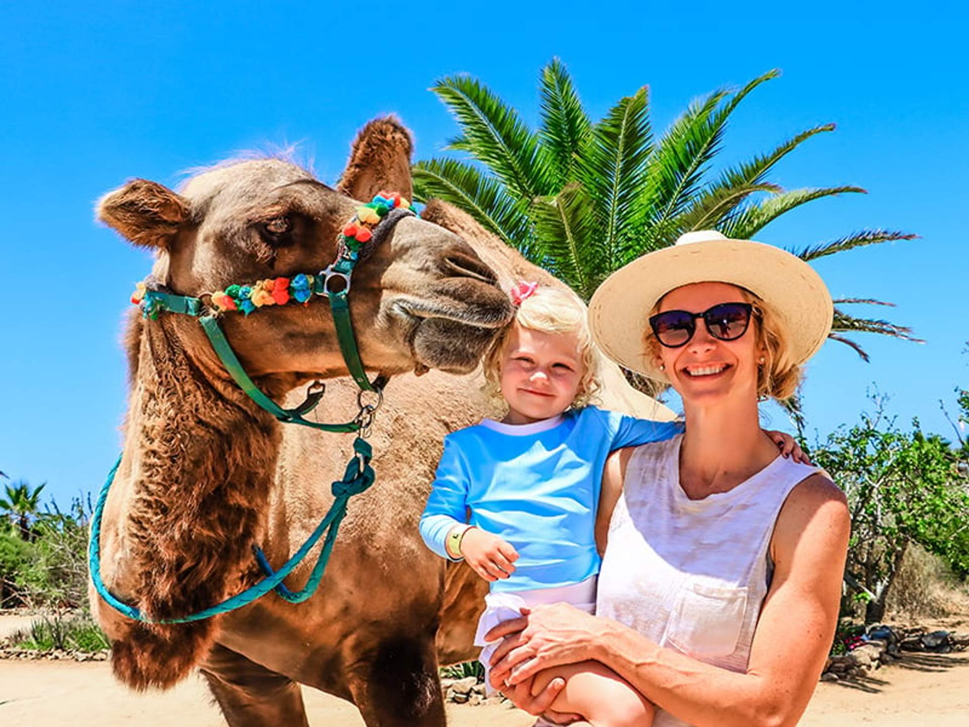 Una mujer y una niña sonríen para una foto con un camello en un día soleado, con palmeras de fondo.