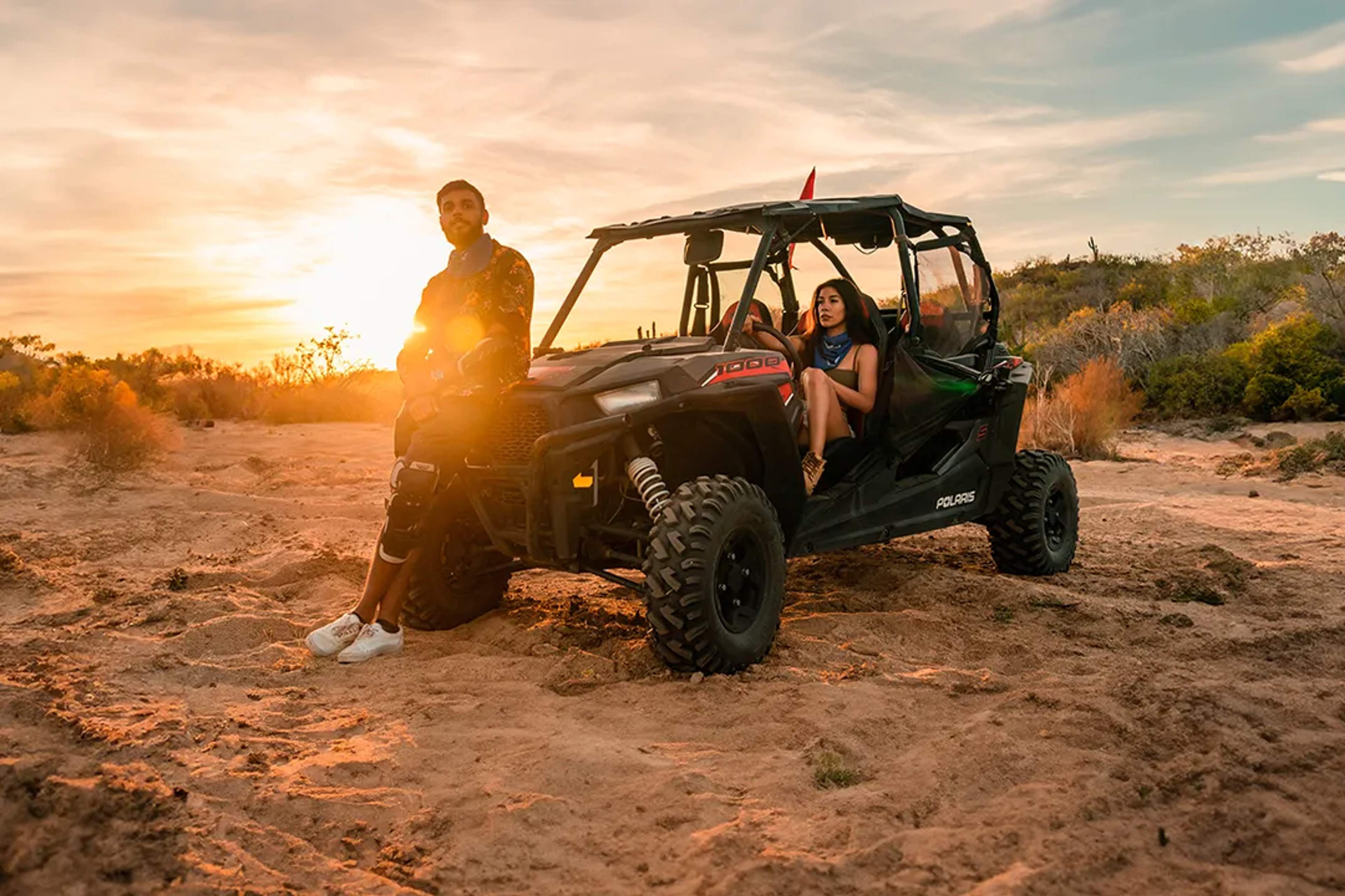 Hombre Y Mujer Con Un Polaris RZR En El Desierto Al Atardecer. Él Está De Pie Y Ella Sentada Dentro Del Vehículo