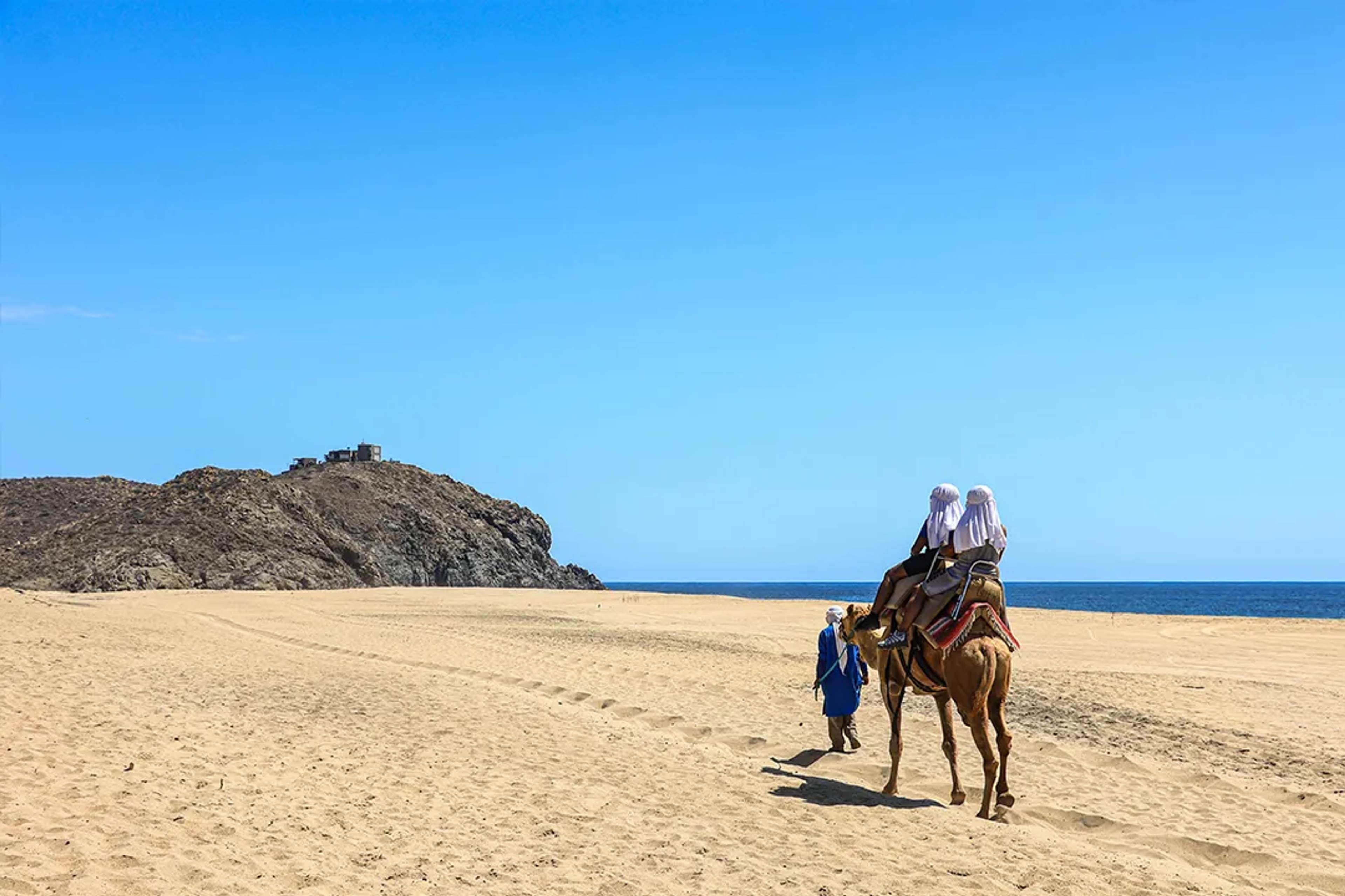 Dos personas montan un camello por una playa desértica con vista al mar y colinas rocosas al fondo.