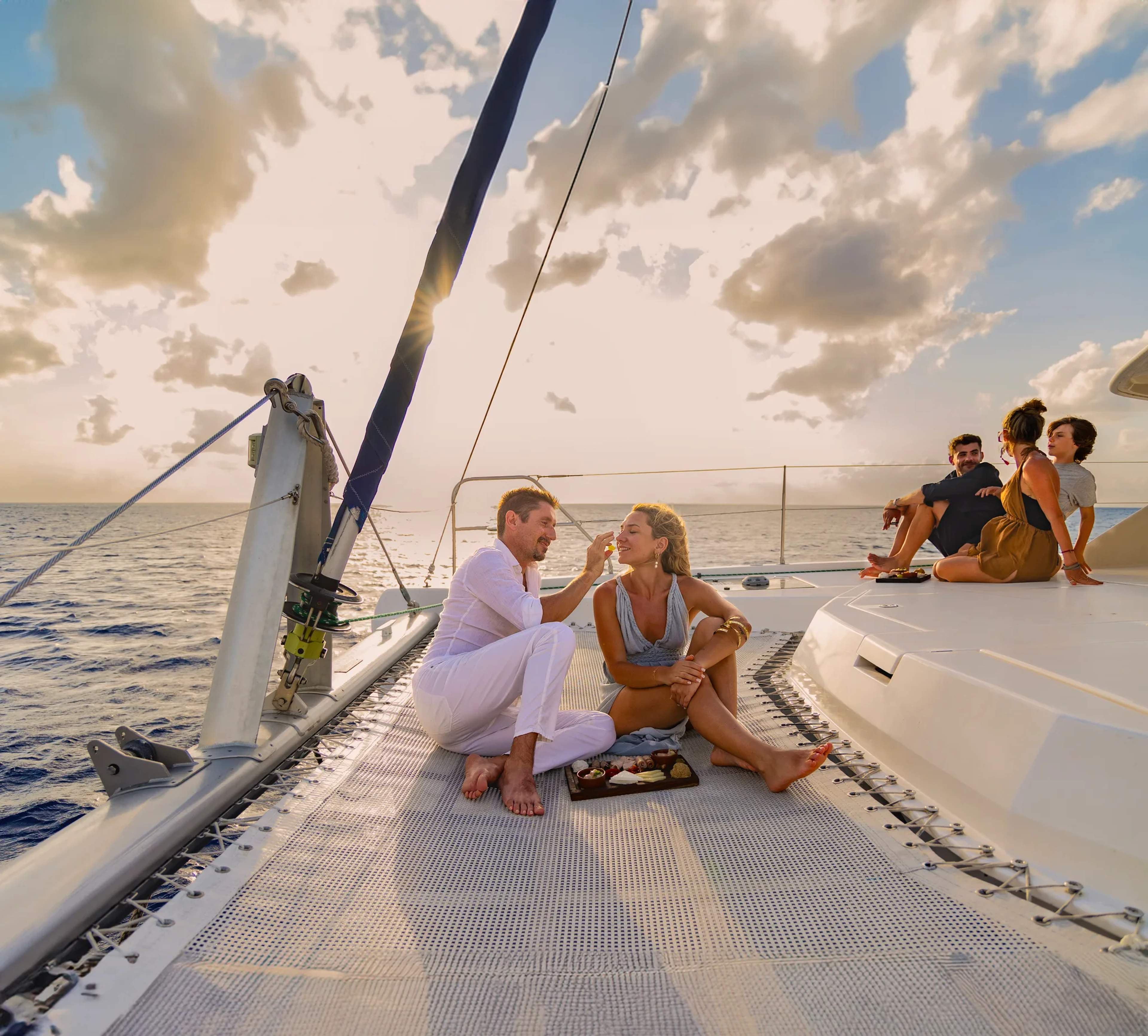 Passengers relax on the deck of a catamaran during a sunset sailing tour, bathed in warm golden-hour light over calm ocean waters.