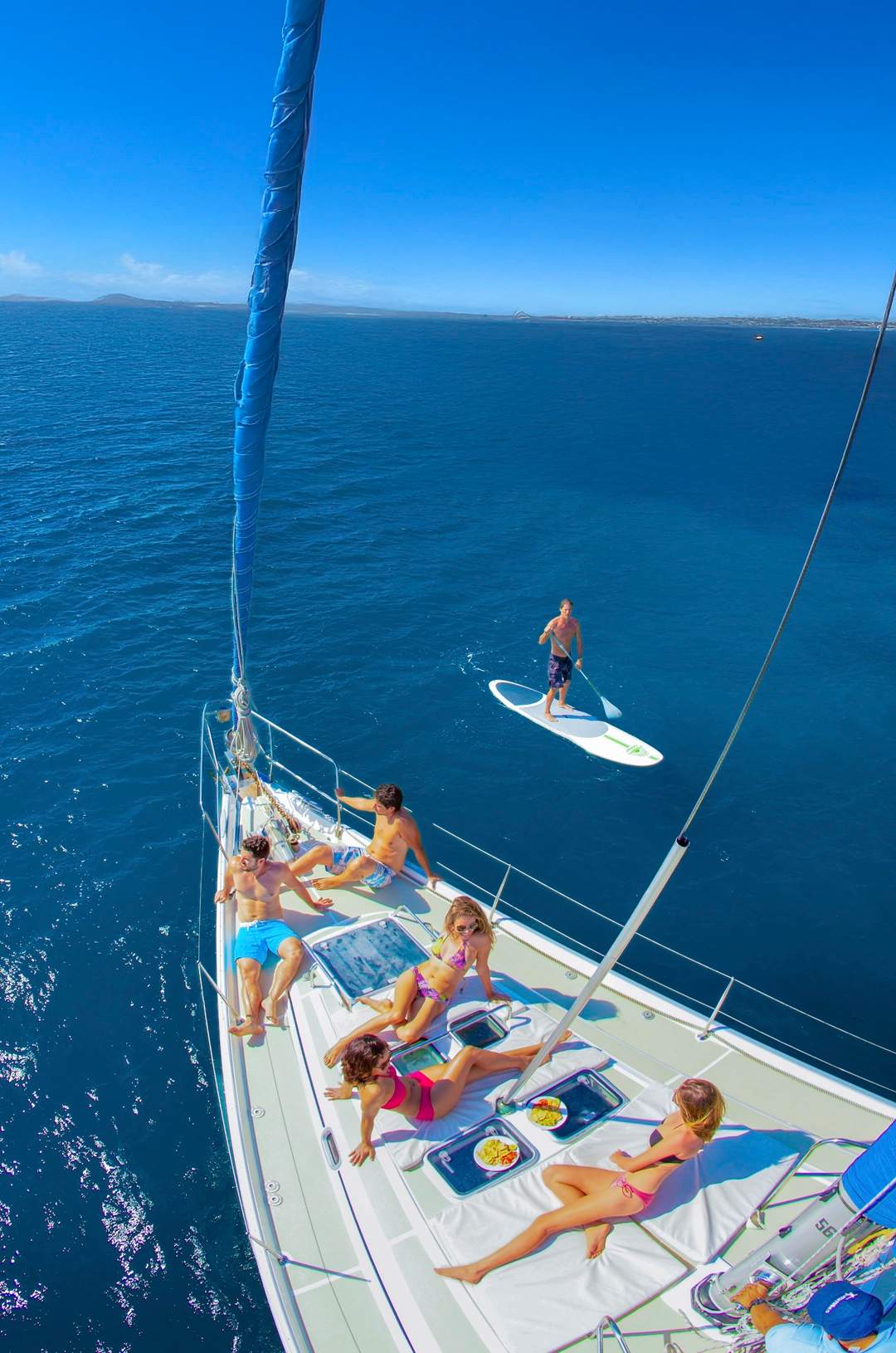 Friends relaxing on a sailboat under a clear blue sky, enjoying the sun and snacks, while one person paddleboards nearby.