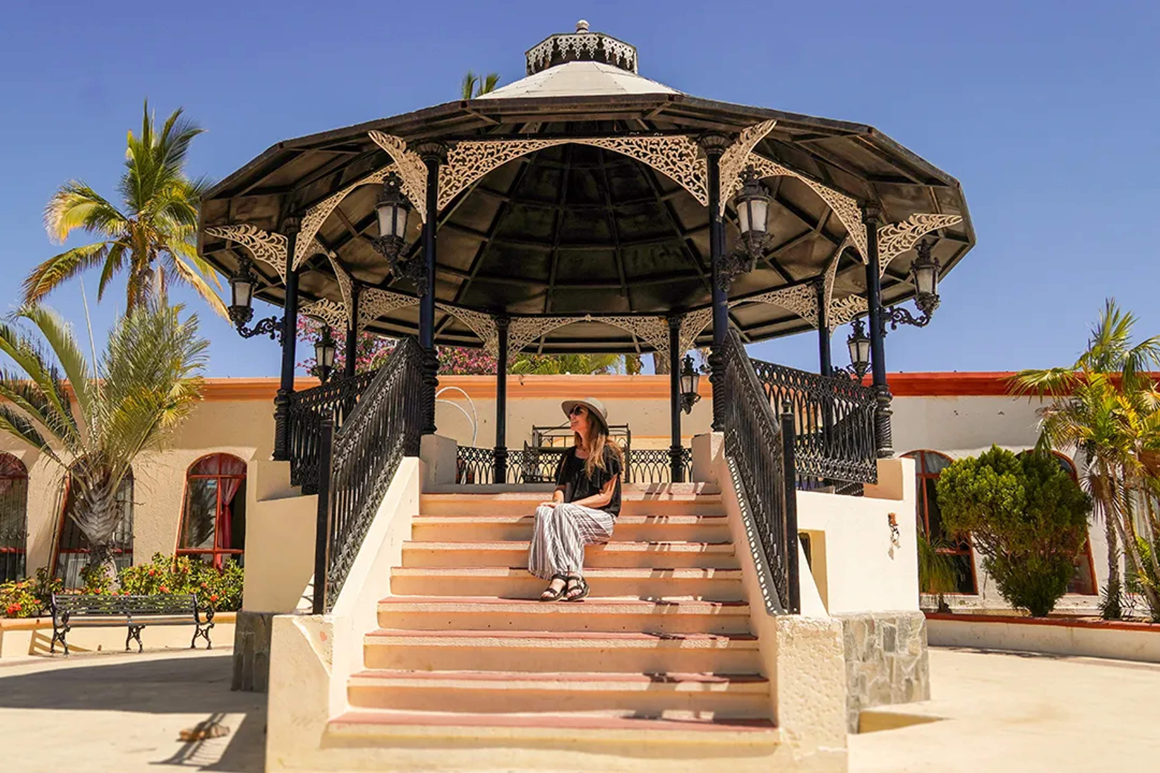 Woman sits on gazebo steps in a sunny colonial-style plaza with palm trees and clear skies.