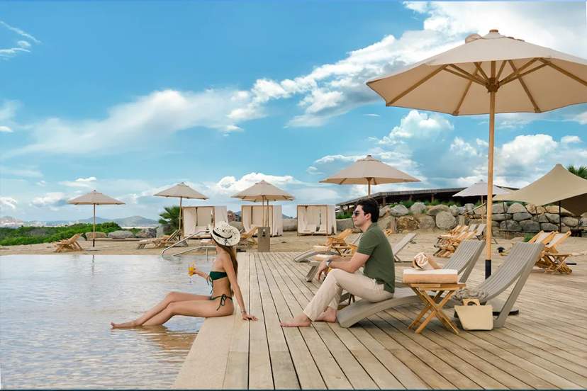 Couple relaxing at beachfront pool lounge in Cabo enjoying tropical drinks under sun and umbrellas.