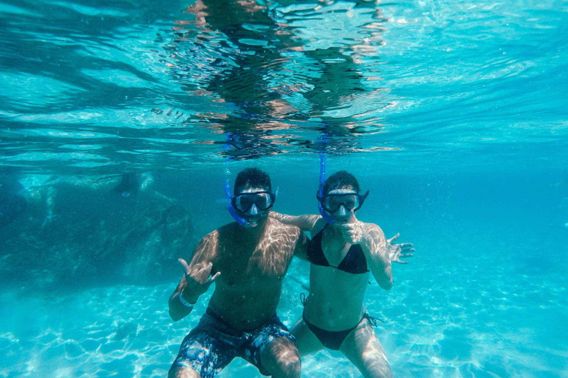 Dos personas haciendo snorkel bajo el agua en aguas azules y claras, usando máscaras de natación y snorkels, posando con gestos de manos.