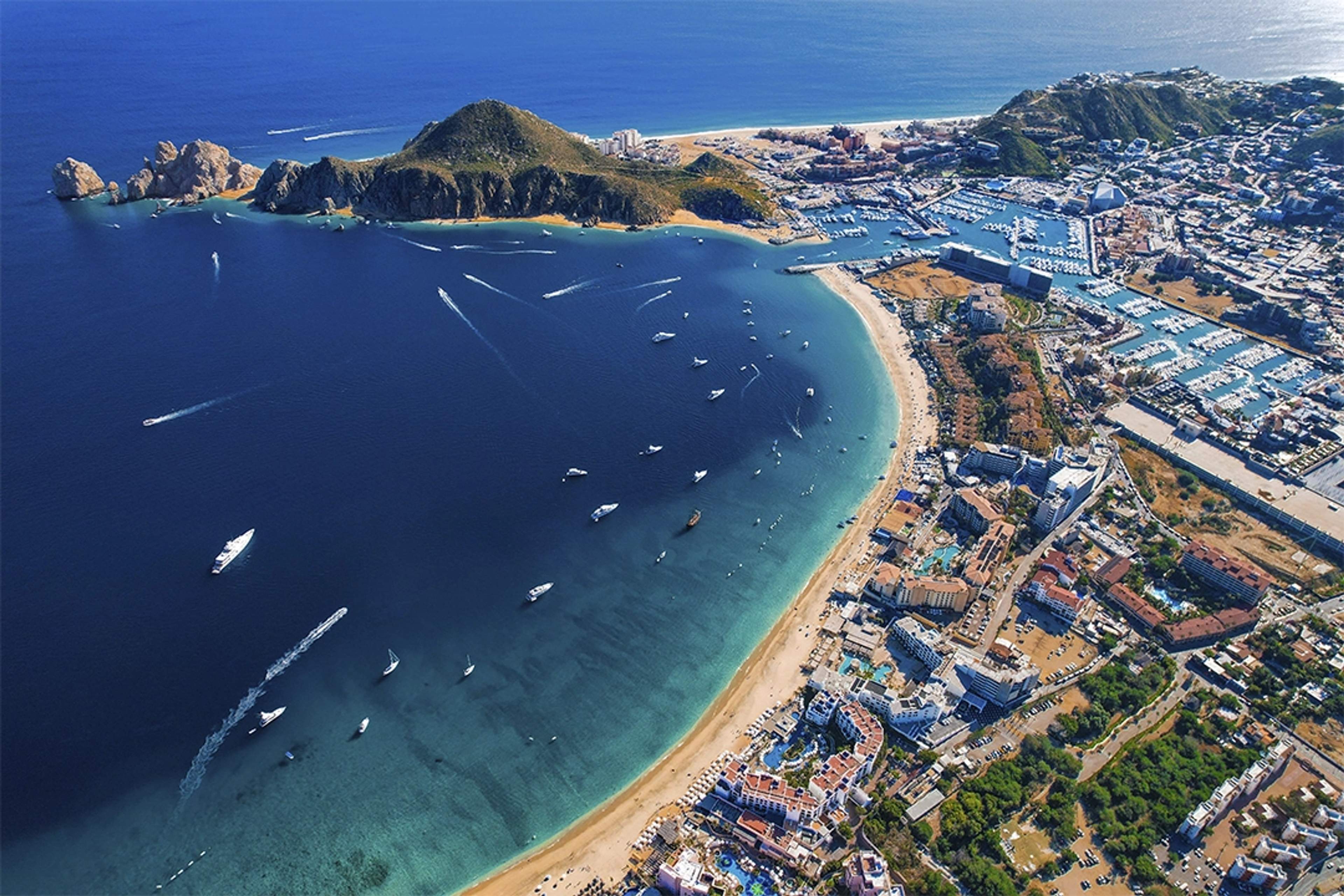 Vista aérea de Cabo San Lucas con botes en la bahía, playas de arena y un animado paisaje urbano costero.