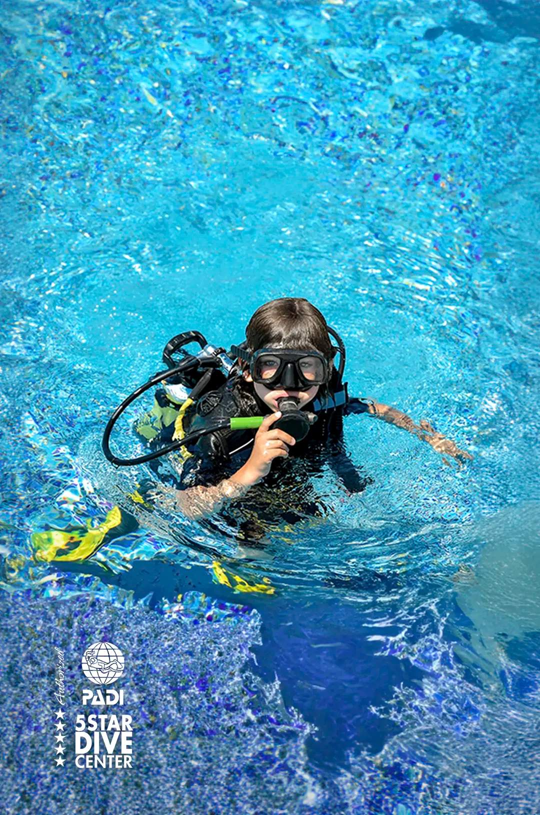 Young scuba diver floating in a pool, practicing with a regulator in clear, sparkling blue water.