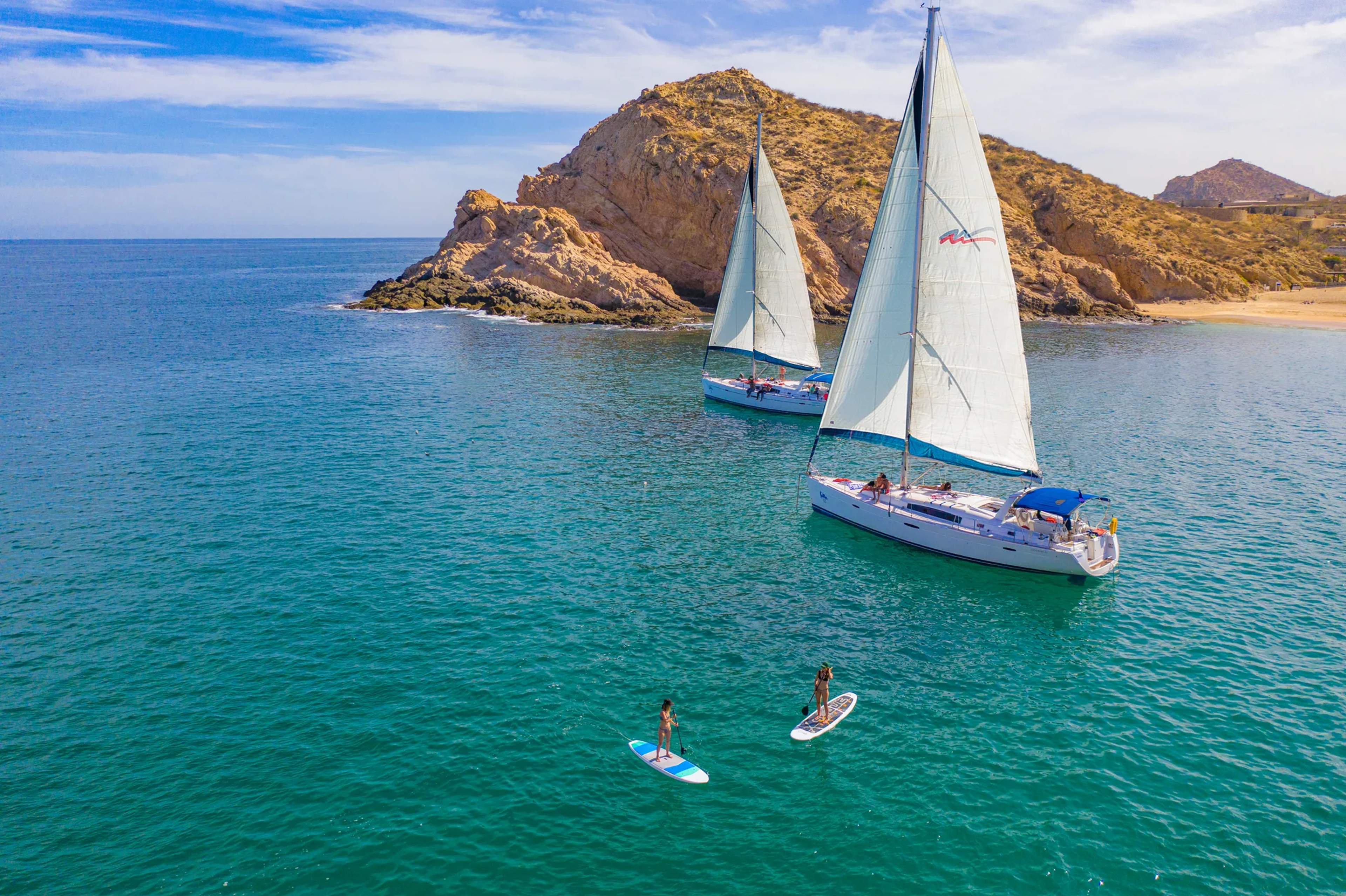 Sailboats anchored in turquoise waters near rocky desert islands while passengers paddleboard in the calm sea.