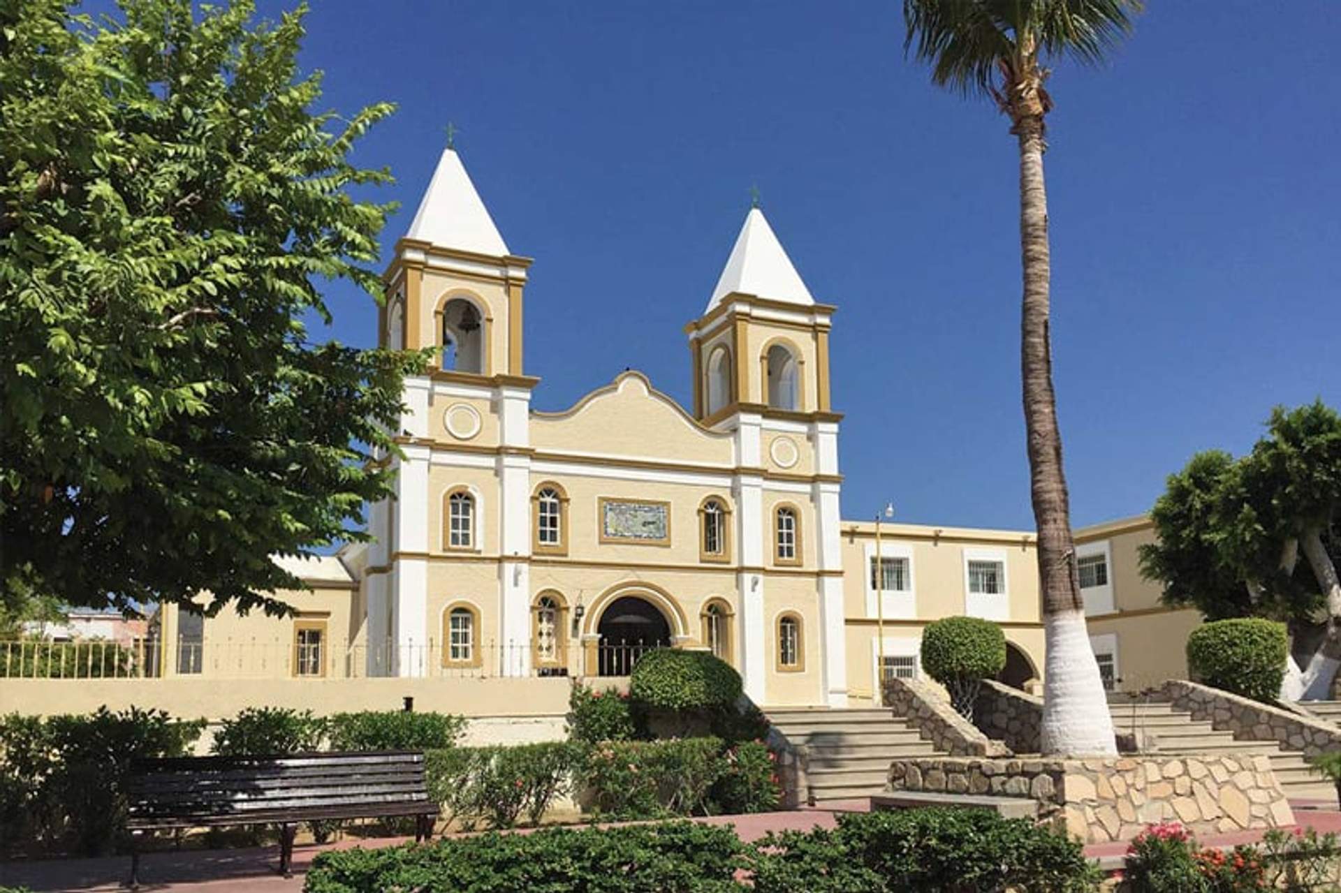 Colonial church with two white bell towers and a yellow facade, surrounded by gardens and trees on a clear day. A tall palm tree is on the right, and there is a park bench in the foreground.