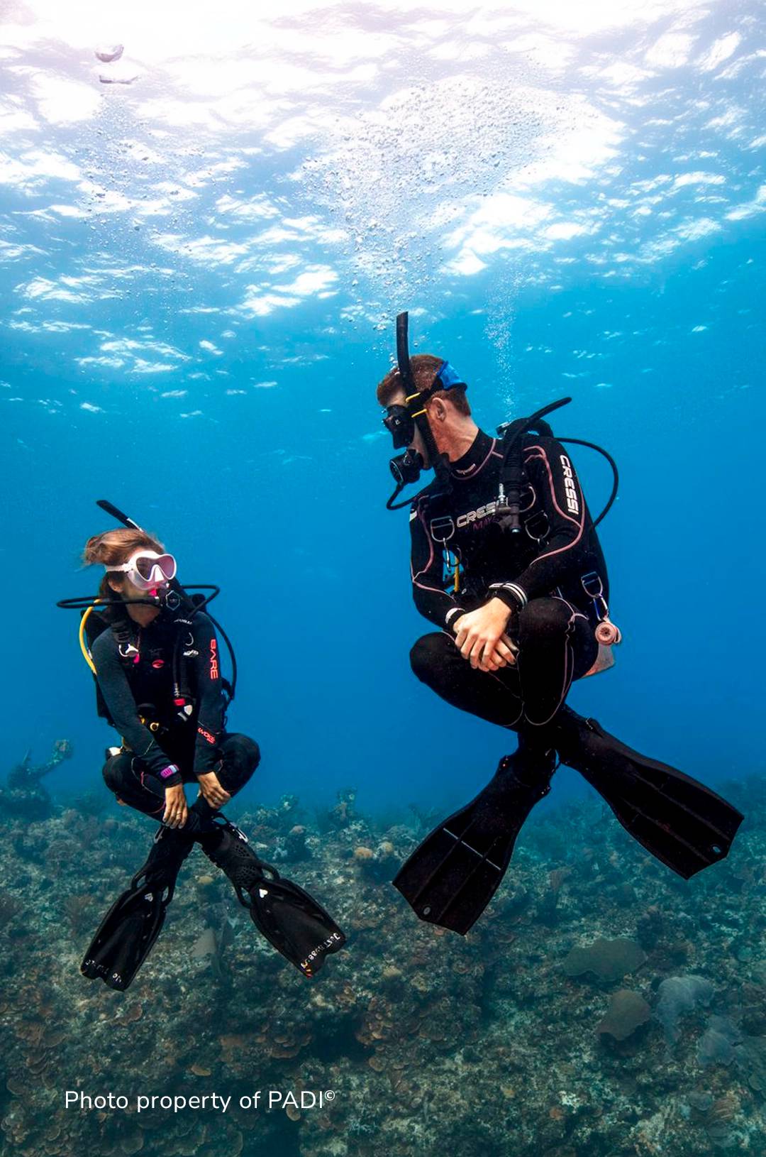  Two scuba divers in black wetsuits hover over a coral reef, interacting calmly as bubbles rise around them.