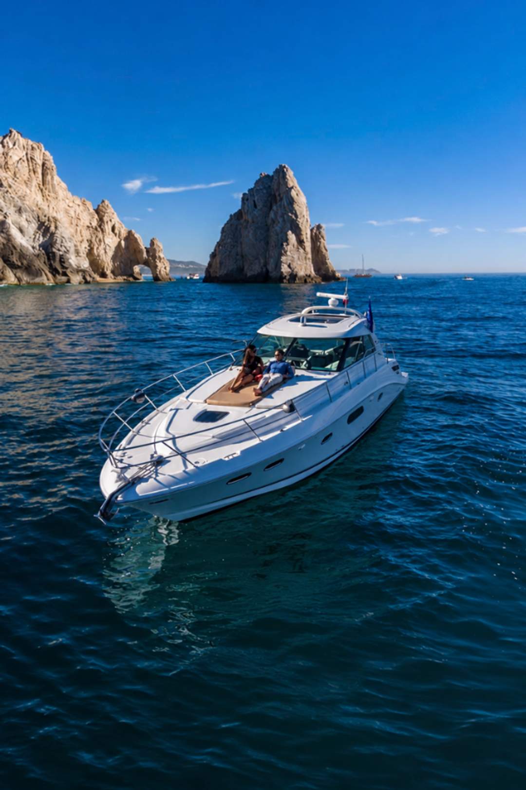 A luxury yacht cruises near the iconic rock formations of Los Cabos under a clear blue sky, with two people relaxing on deck