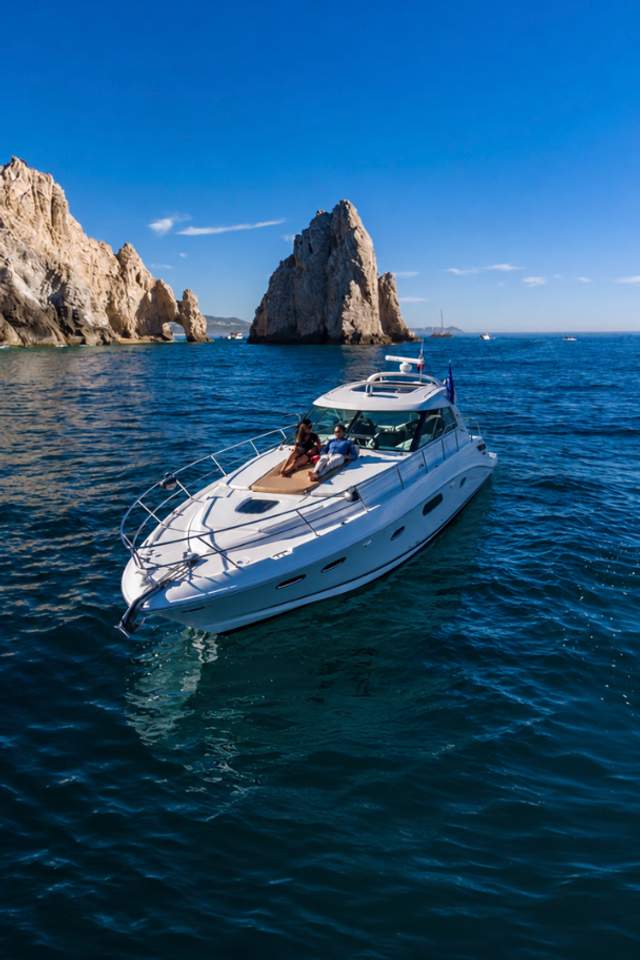  A luxury yacht cruises near the iconic rock formations of Los Cabos under a clear blue sky, with two people relaxing on deck