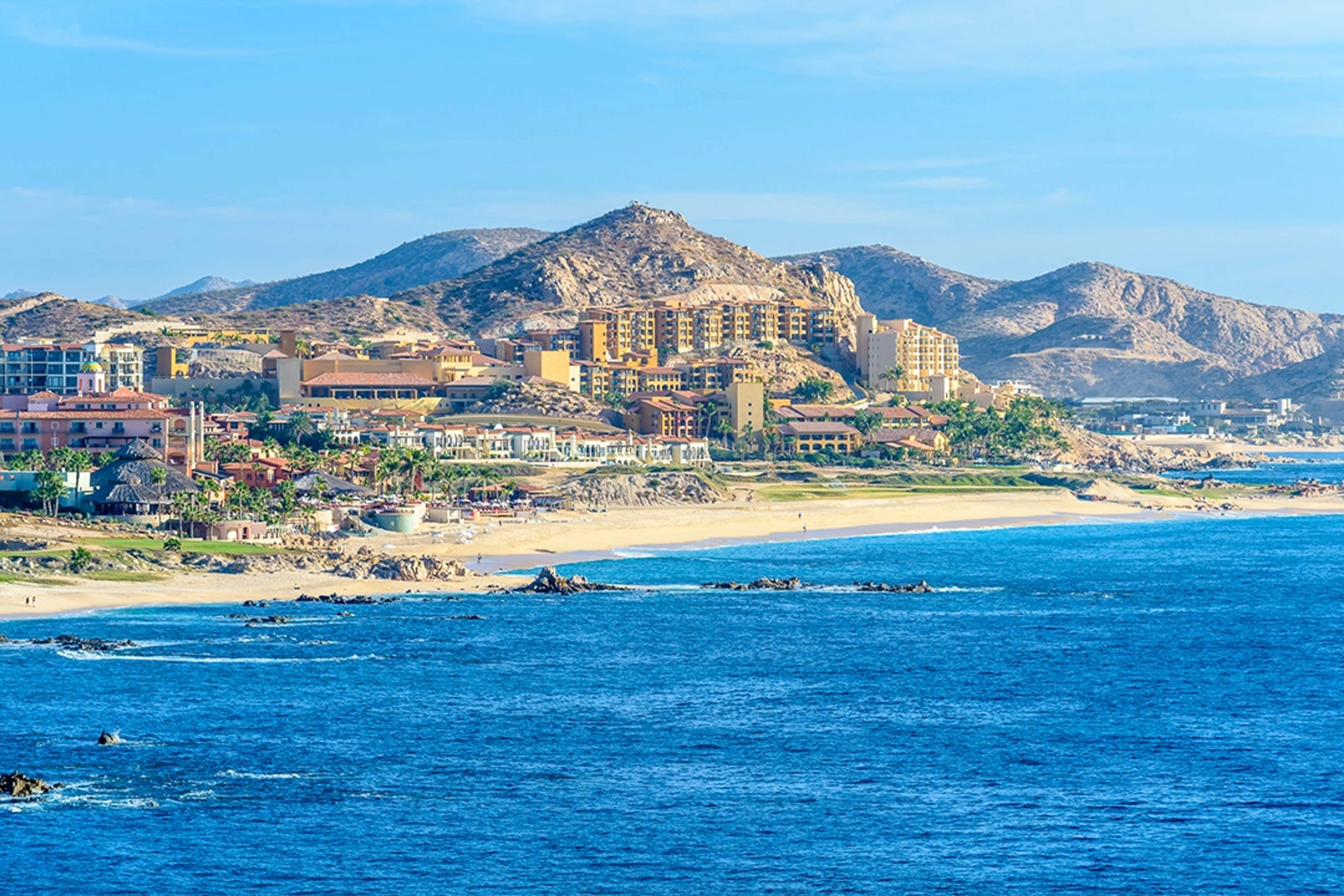 Vista panorámica de los resorts de Cabo San Lucas, situados a lo largo de la costa, rodeados de montañas y el mar azul profundo.