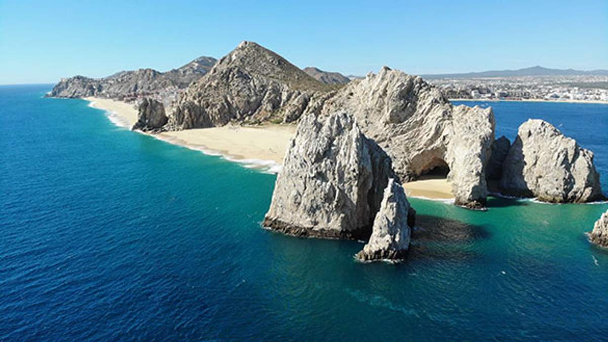 A breathtaking aerial view of the iconic rock formations and pristine beaches of Cabo San Lucas, Mexico, where the deep blue sea meets the golden sandy shoreline under a clear sky.