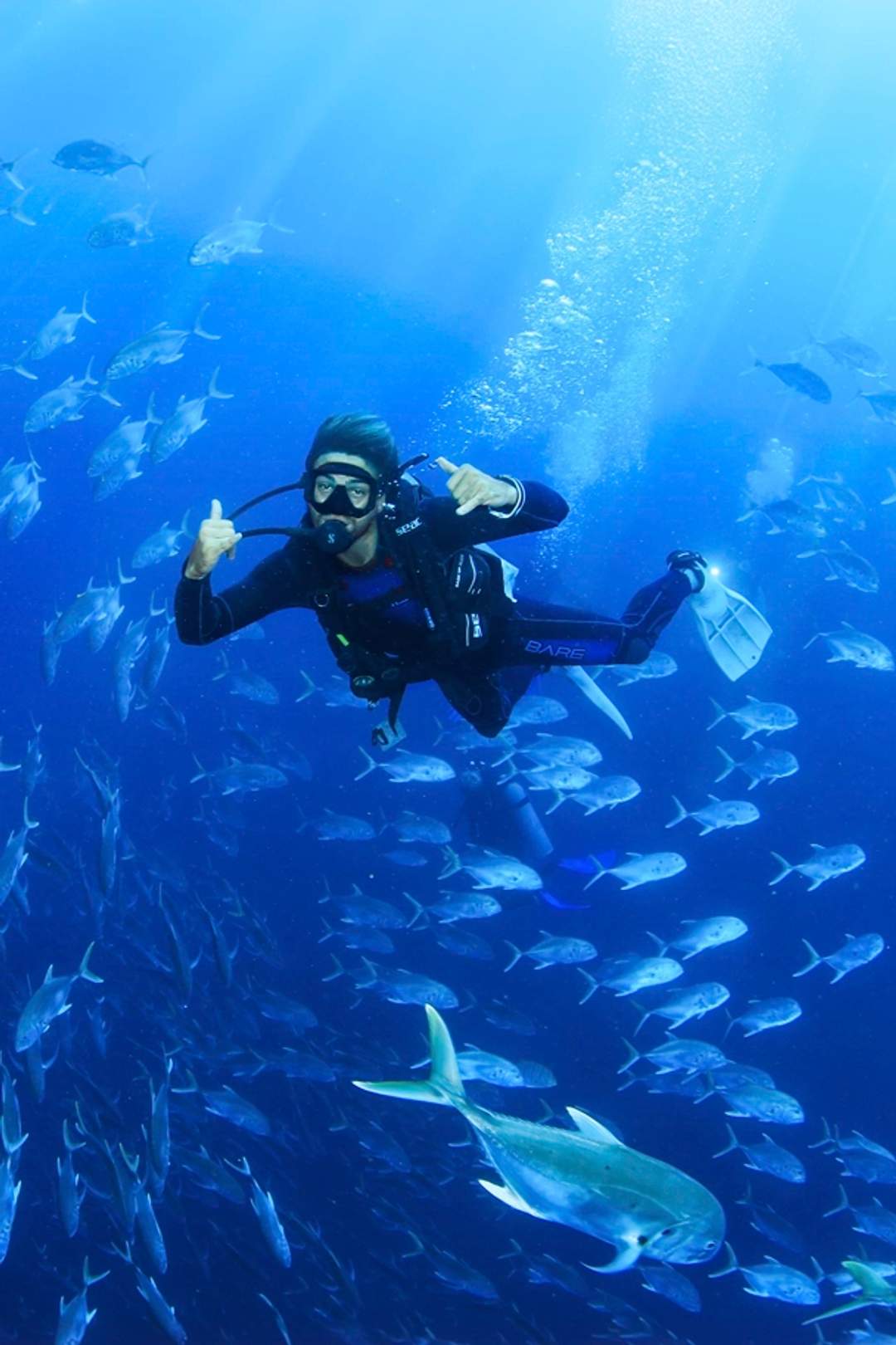 Un buzo rodeado de un banco de peces, haciendo una señal de aprobación, con la luz del sol filtrándose a través del agua.