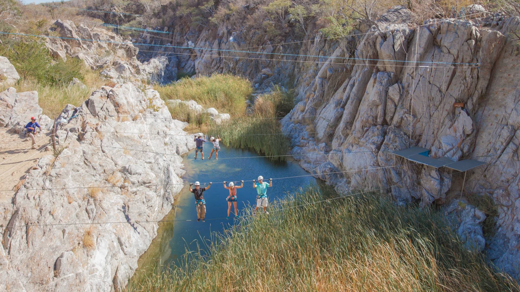Personas cruzando un puente colgante estrecho sobre un desfiladero con una pequeña piscina abajo, usando cascos y arneses.