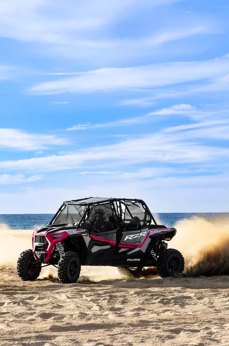 Un UTV levanta arena mientras avanza rápido por la playa, con el océano de fondo bajo un cielo parcialmente nublado.