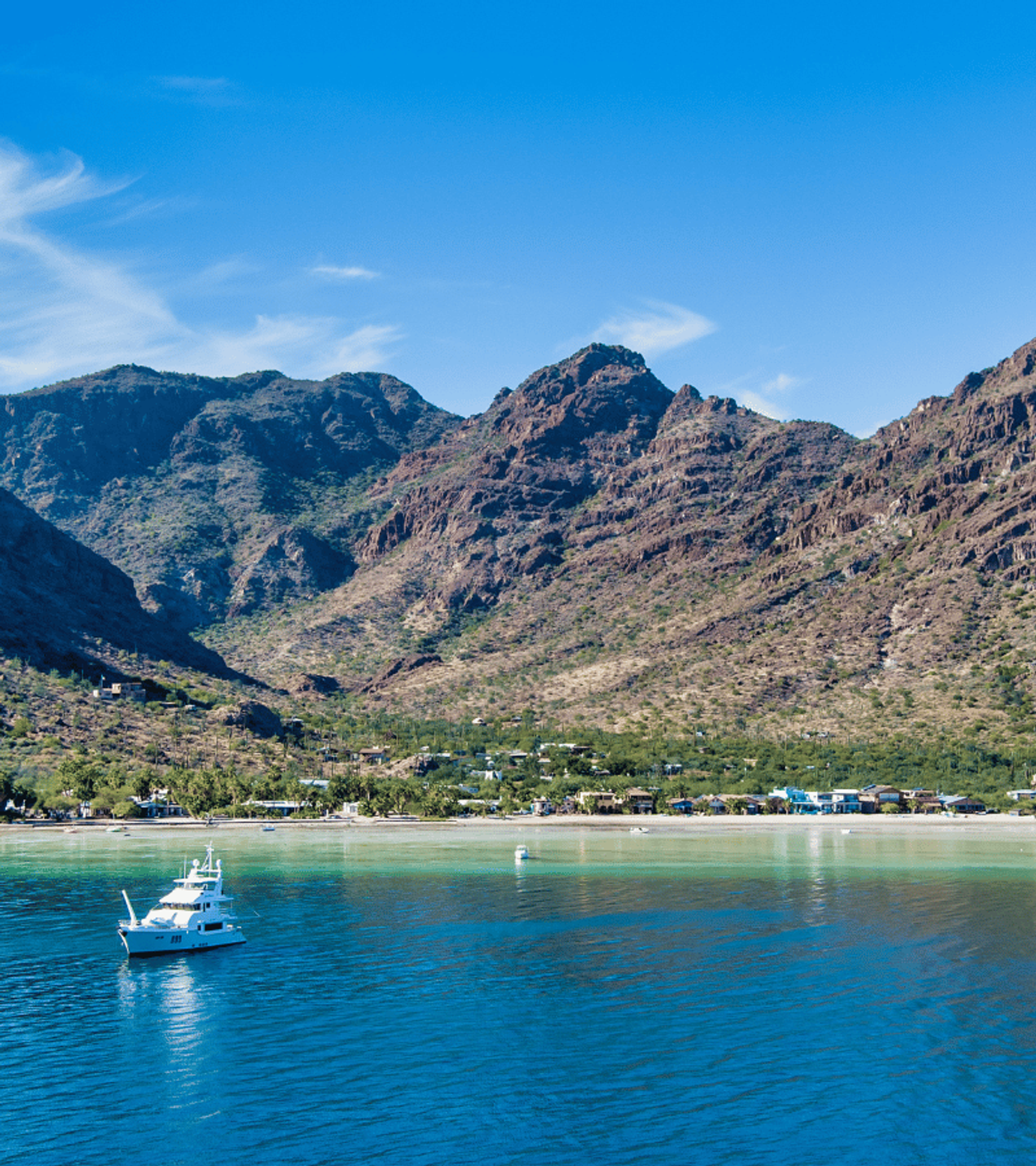 Vista de una costa con un yate en aguas azules, una playa bordeada de casas y vegetación, y colinas rocosas bajo un cielo azul claro.