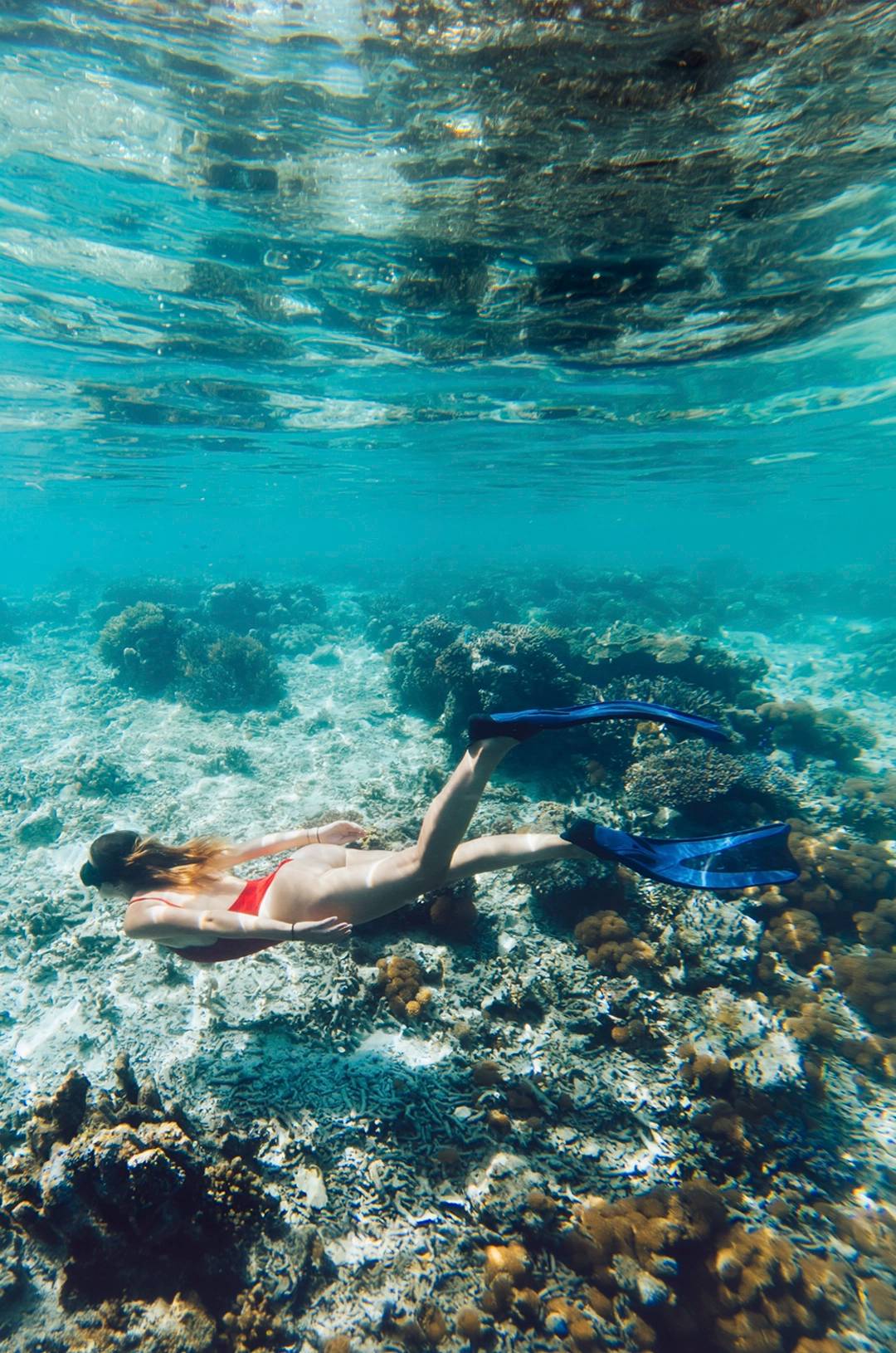 A snorkeler in a red swimsuit exploring a coral reef underwater, wearing fins in clear blue water.