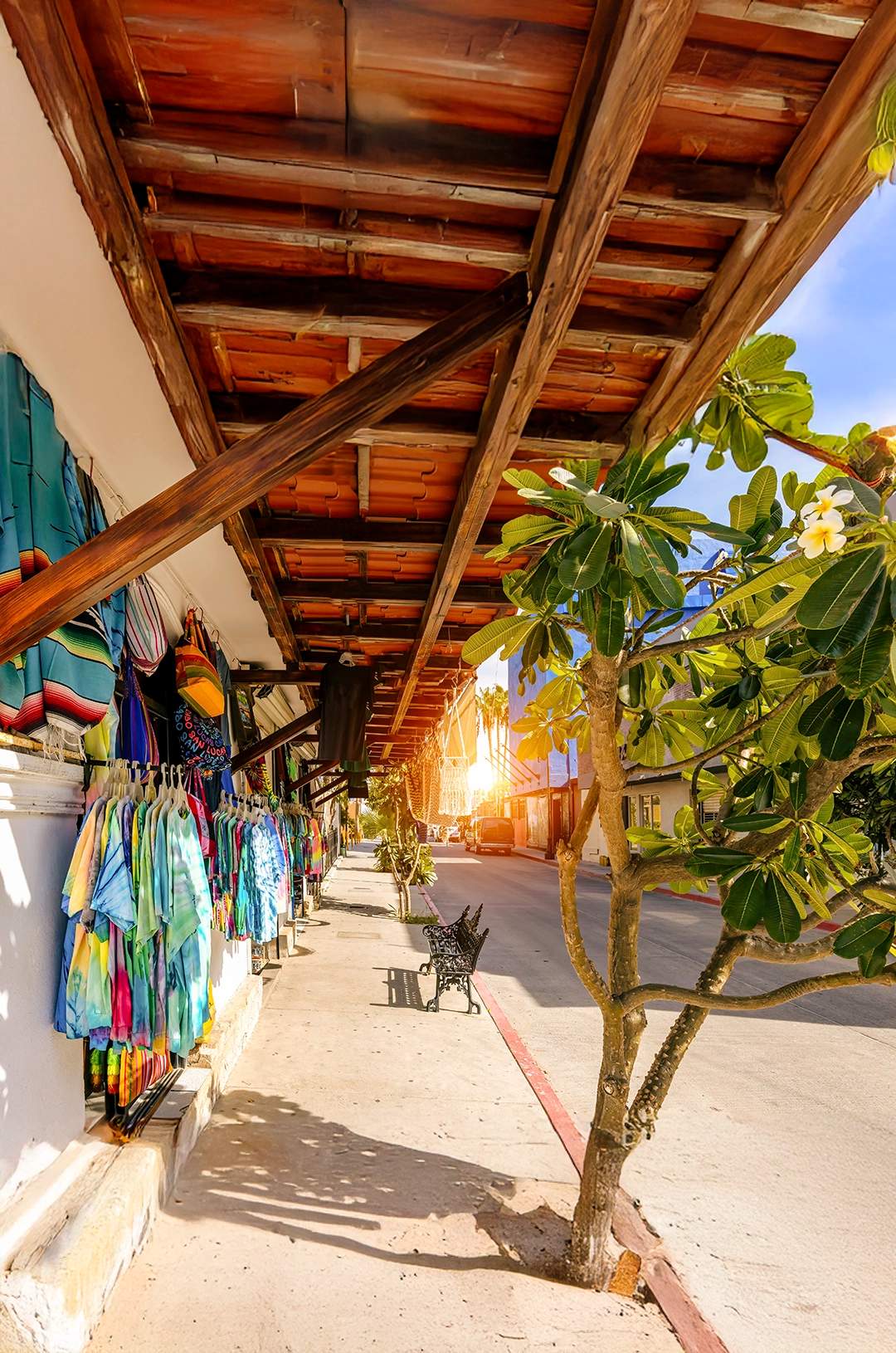 A charming street in Cabo San Lucas with colorful clothing shops, wooden roofs, and a golden sunset.