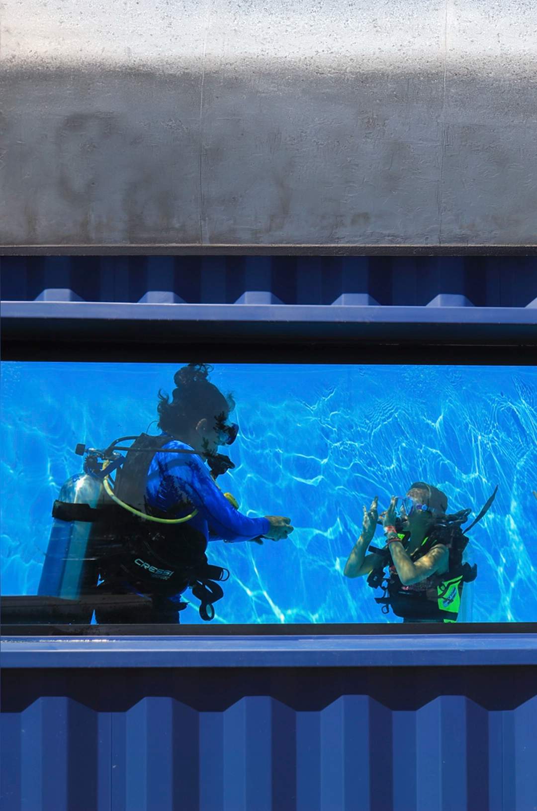 Scuba divers in a training pool practice breathing techniques, seen through a window with "#CaboAdventures" on it.