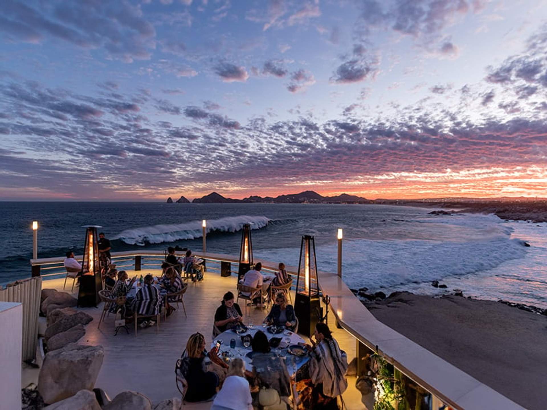 Personas cenando en una terraza al aire libre con vista al océano al atardecer, con nubes dramáticas en el cielo.
