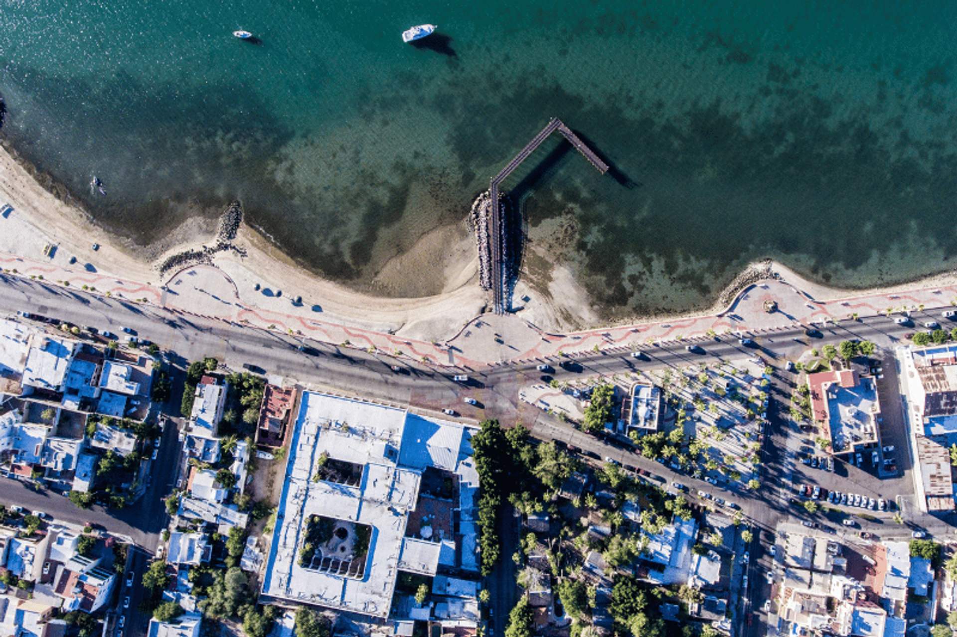 Vista aérea del malecón de La Paz, mostrando playas de arena, un muelle, barcos en el agua y edificios bordeando la costa.