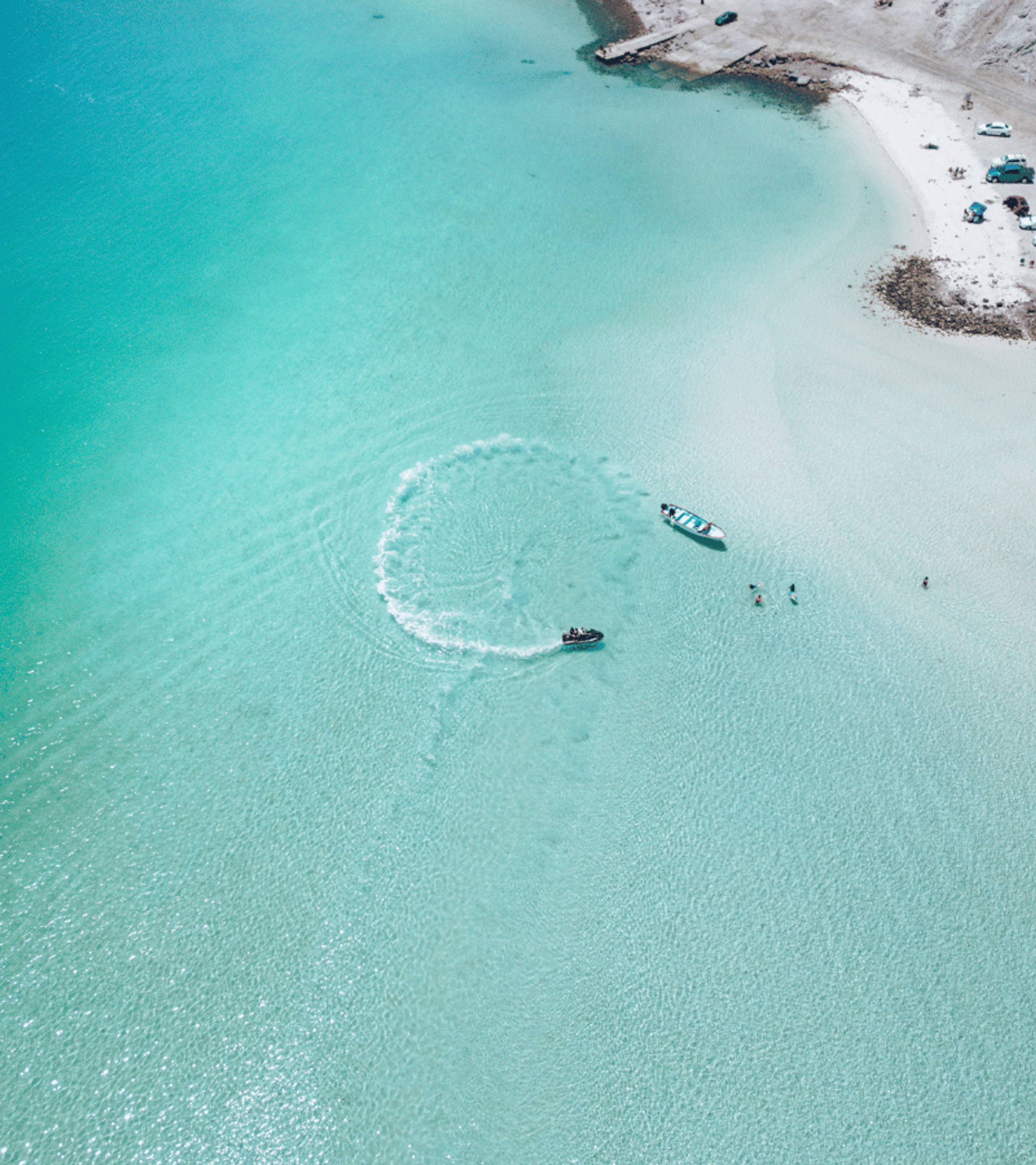 Moto acuática trazando un círculo en el agua turquesa junto a una playa con arena blanca y algunas personas en la orilla