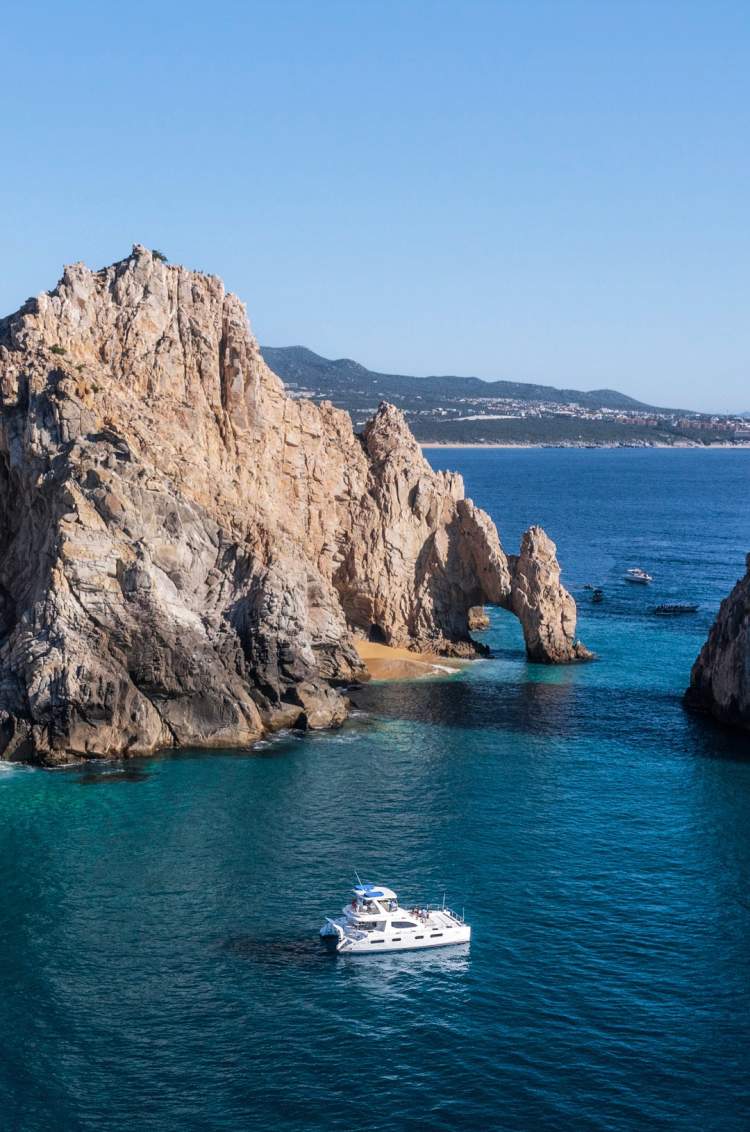  A boat sailing near the dramatic rock formations of Cabo San Lucas with clear blue water and mountains in the background.