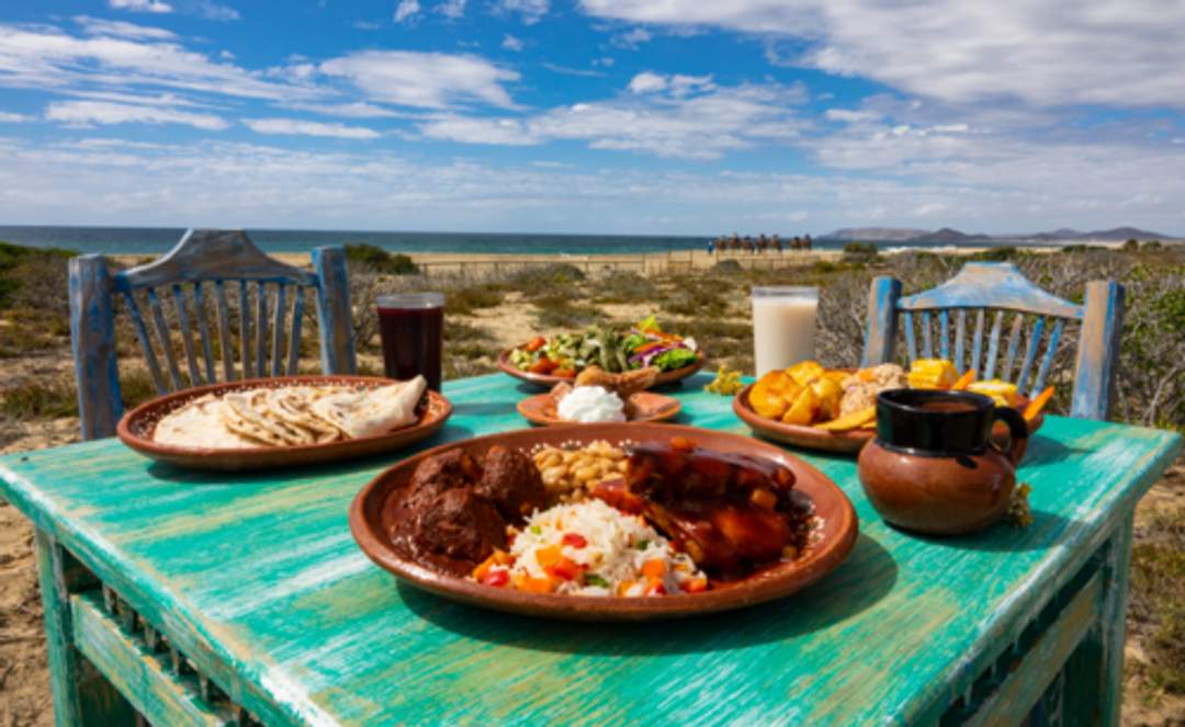 Una mesa colorida llena de platillos tradicionales mexicanos, ubicada en una playa con vista al mar en Cabo San Lucas.