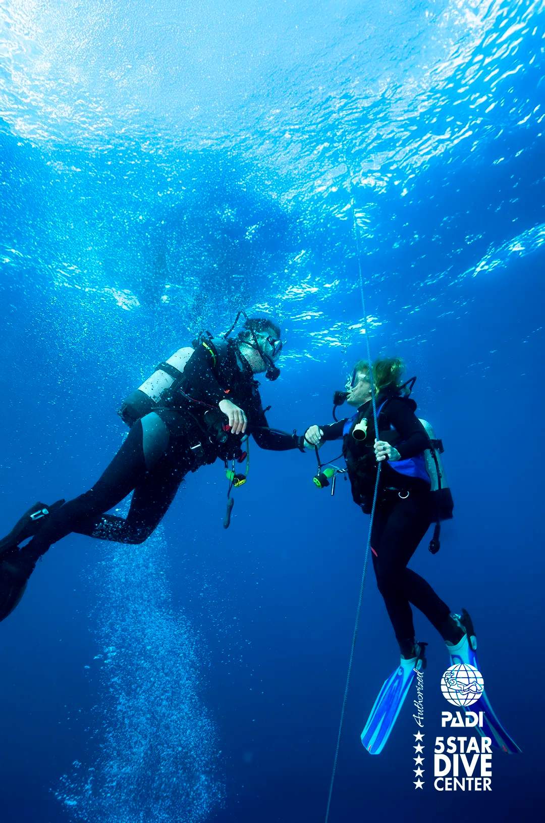 Two scuba divers hold onto a guide rope underwater, communicating as bubbles rise around them in the deep blue ocean.