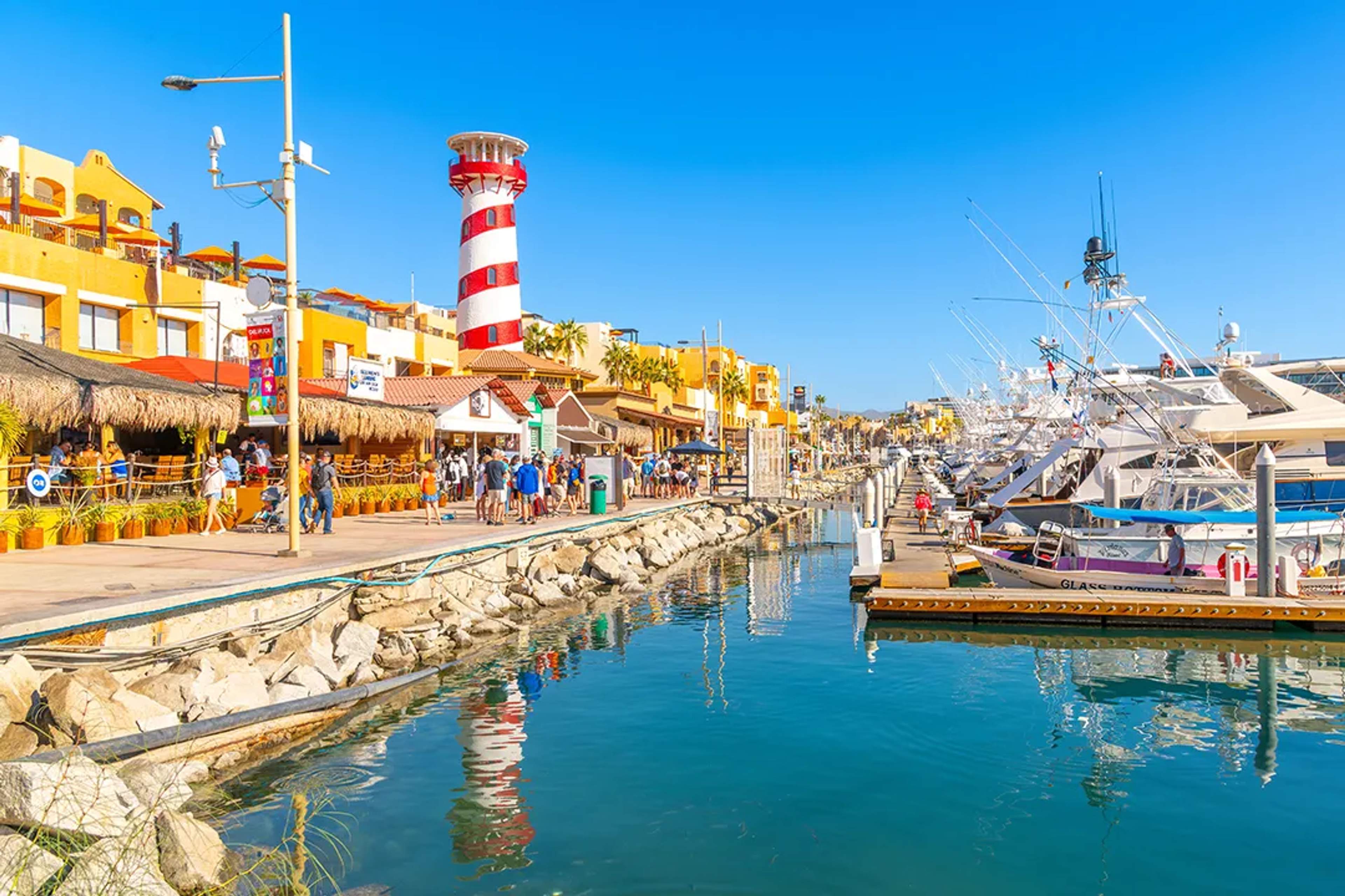 Marina Cabo San Lucas waterfront with lighthouse, shops and yachts on a sunny day