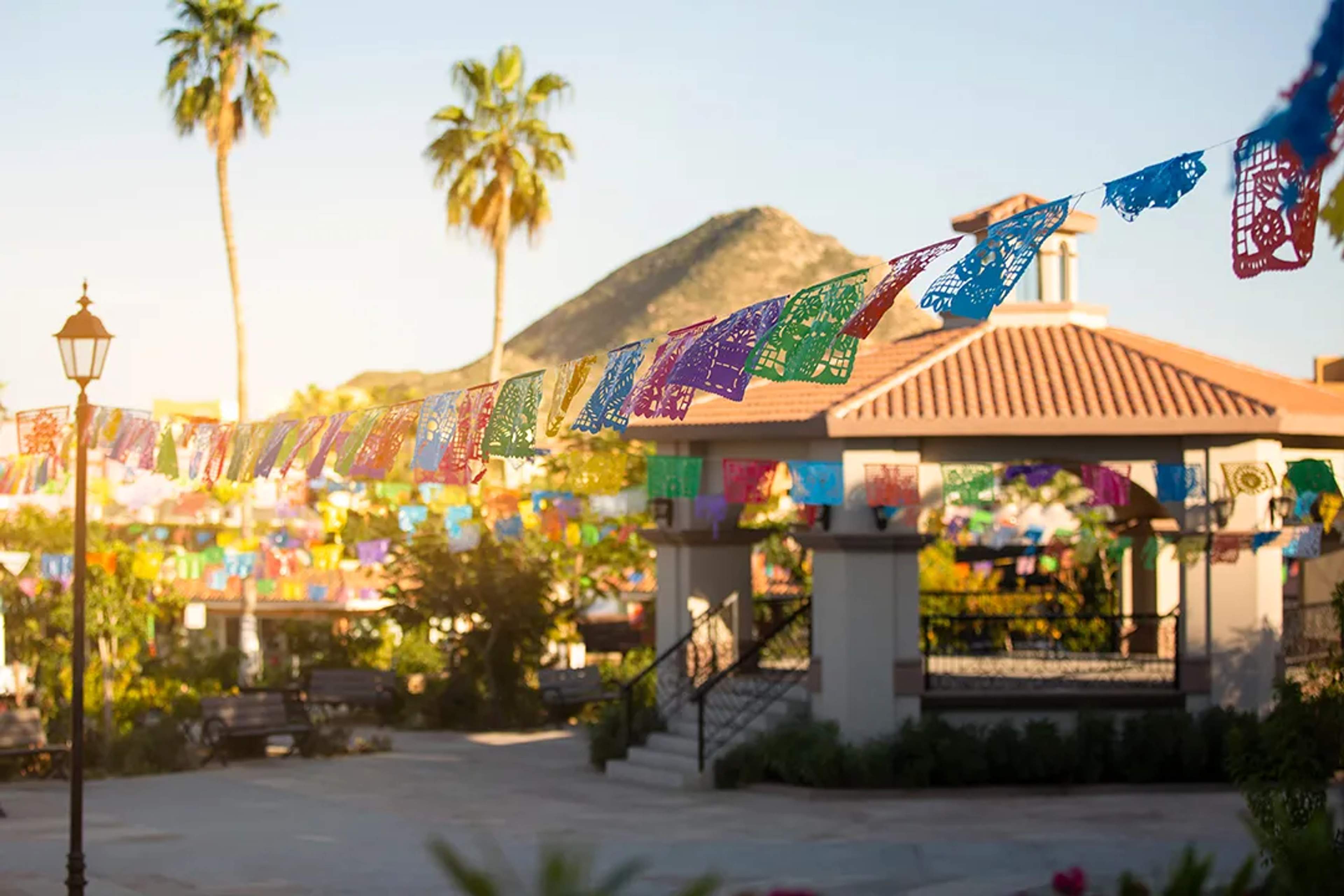 Colorful papel picado decorating a sunny Baja plaza with palm trees and mountain views.